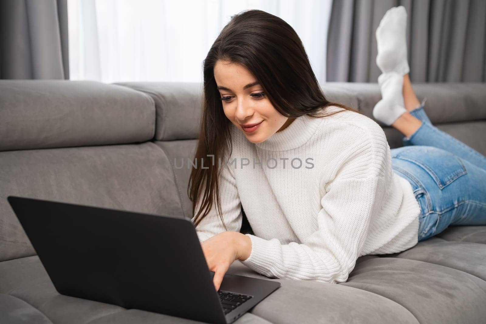 Young female laying on the sofa and using her notebook. A smiling woman has online shopping using a laptop at home. Working from home as a freelancer.