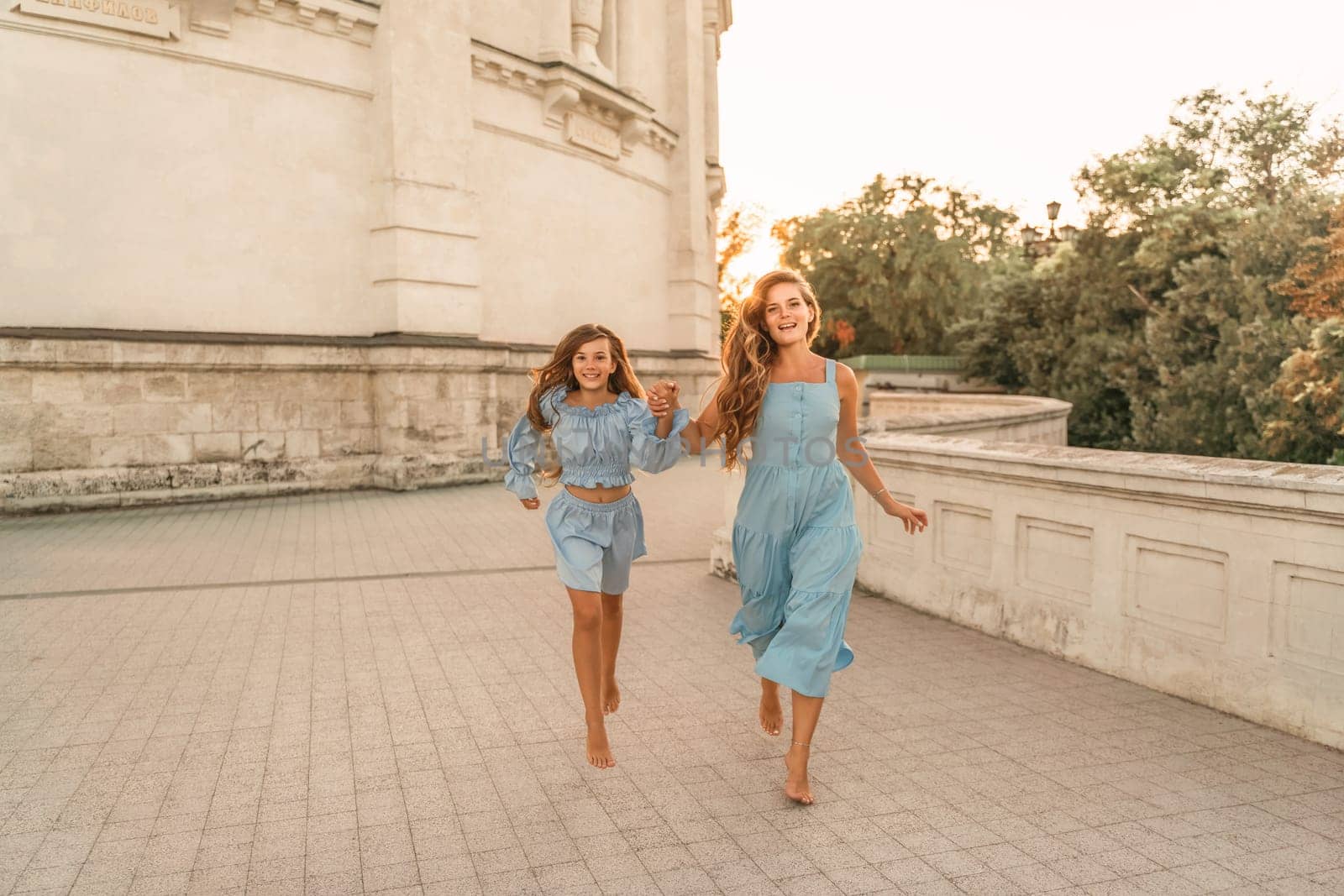 Daughter mother run holding hands. In blue dresses with flowing long hair against the backdrop of a sunset and a white building. by Matiunina