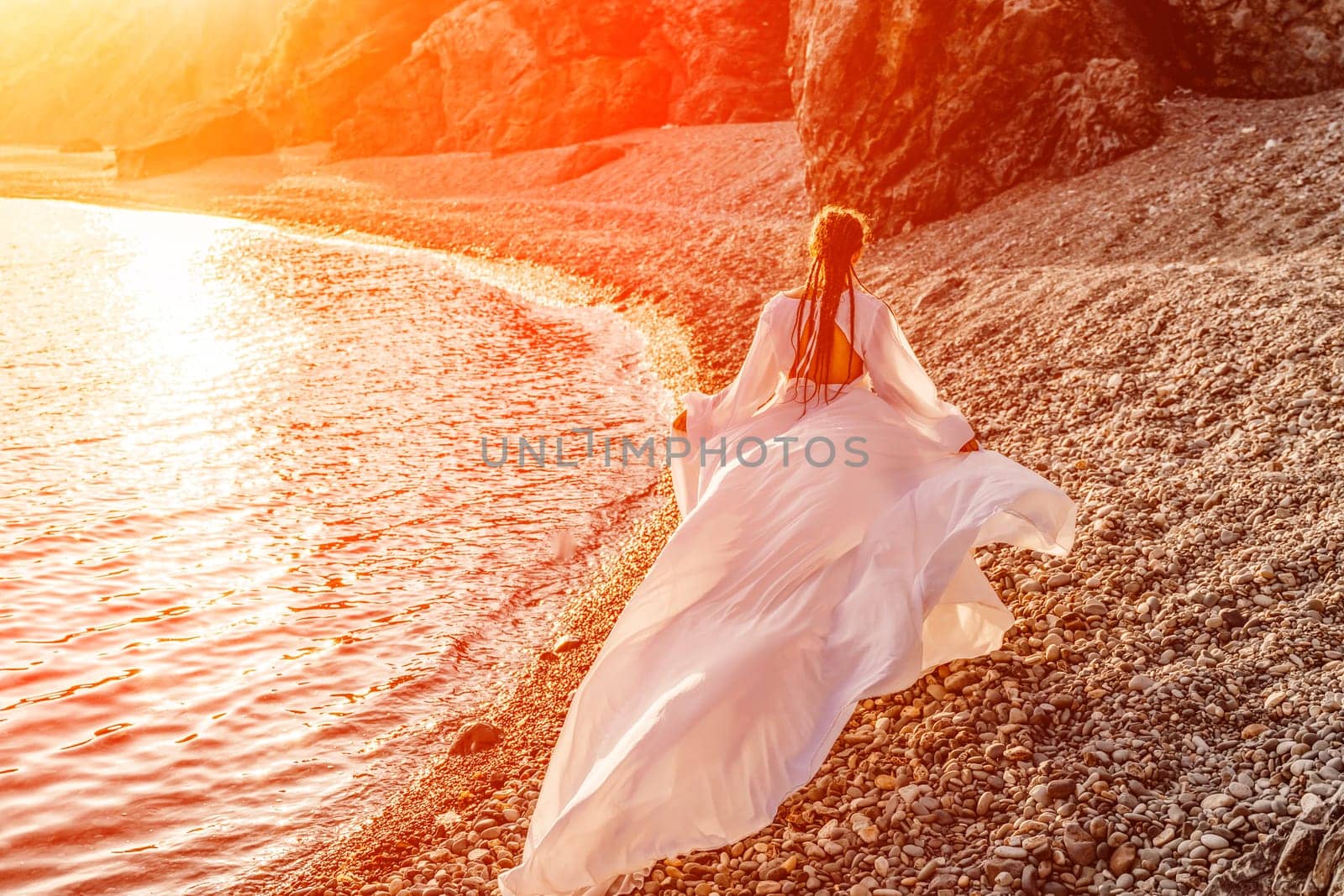 Mysterious woman silhouette long hair walks on the beach ocean water, sea nymph wind listens to the wave. Throws up a long white dress, a divine sunset. Artistic photo from the back without a face.