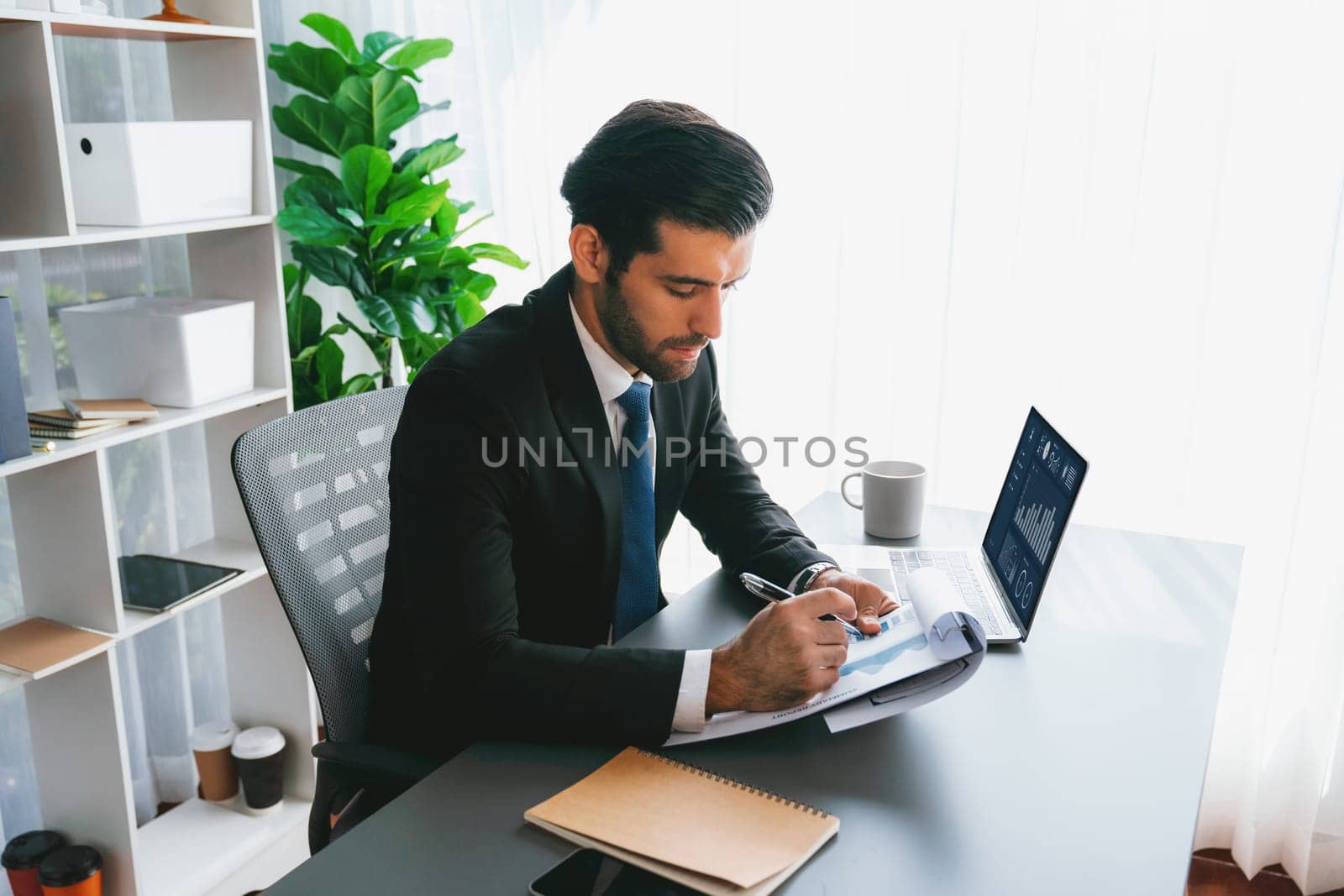 Modern professional businessman at modern office desk using laptop to work and write notes. Diligent office worker working on computer notebook in his office work space. fervent