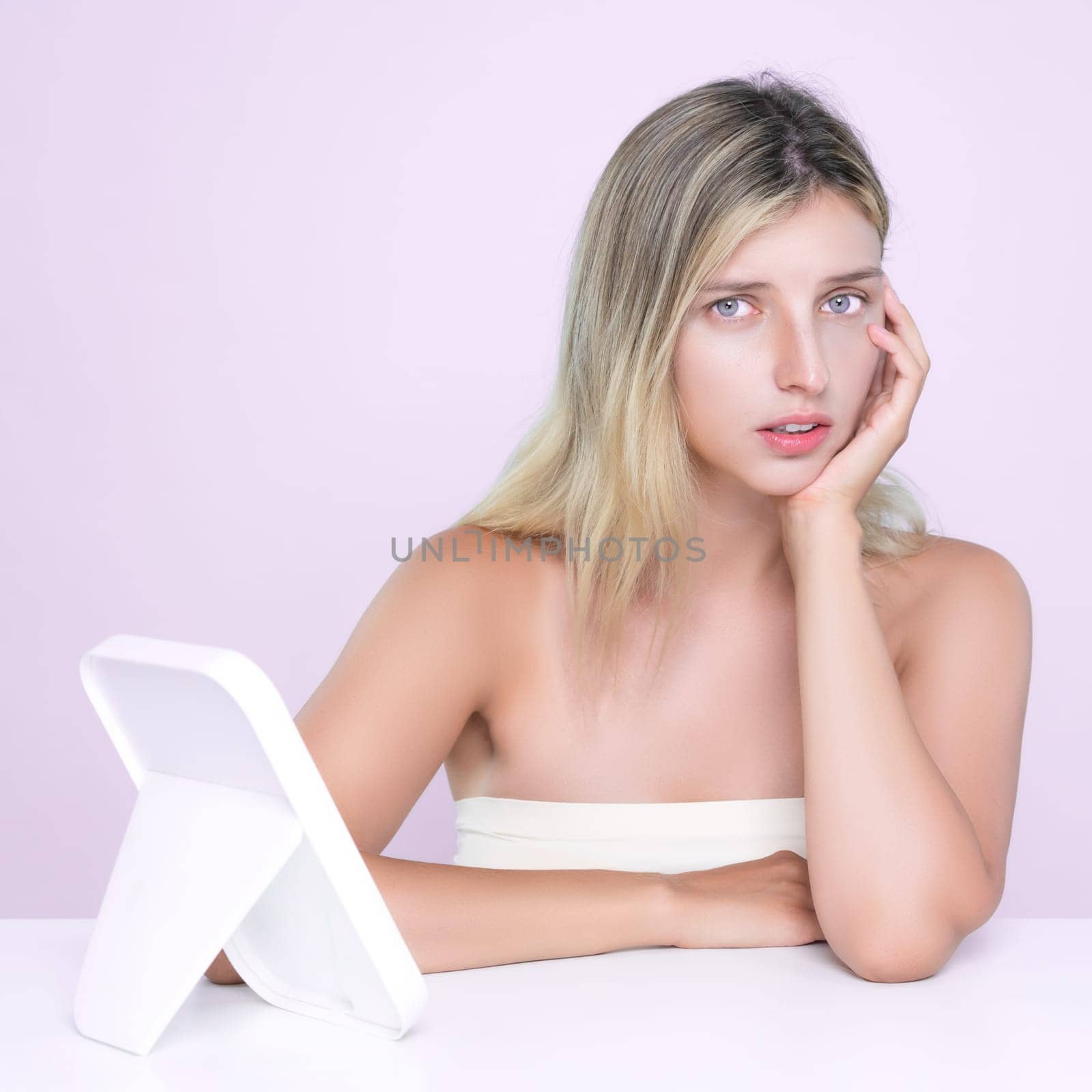 Alluring portrait of beautiful woman feel insecure on her facial skin, oily or acne problem. Worried female model with perfect smooth clean skin check her face on mirror in pink isolated background.