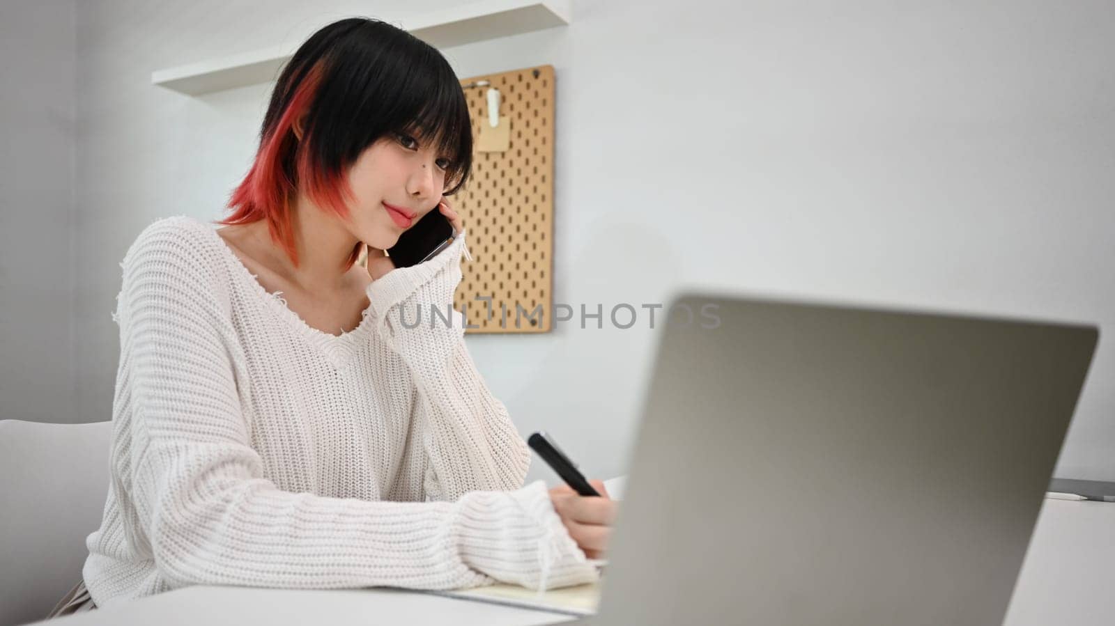 Beautiful female small business owner looking at laptop screen, communicating with client on mobile phone by prathanchorruangsak