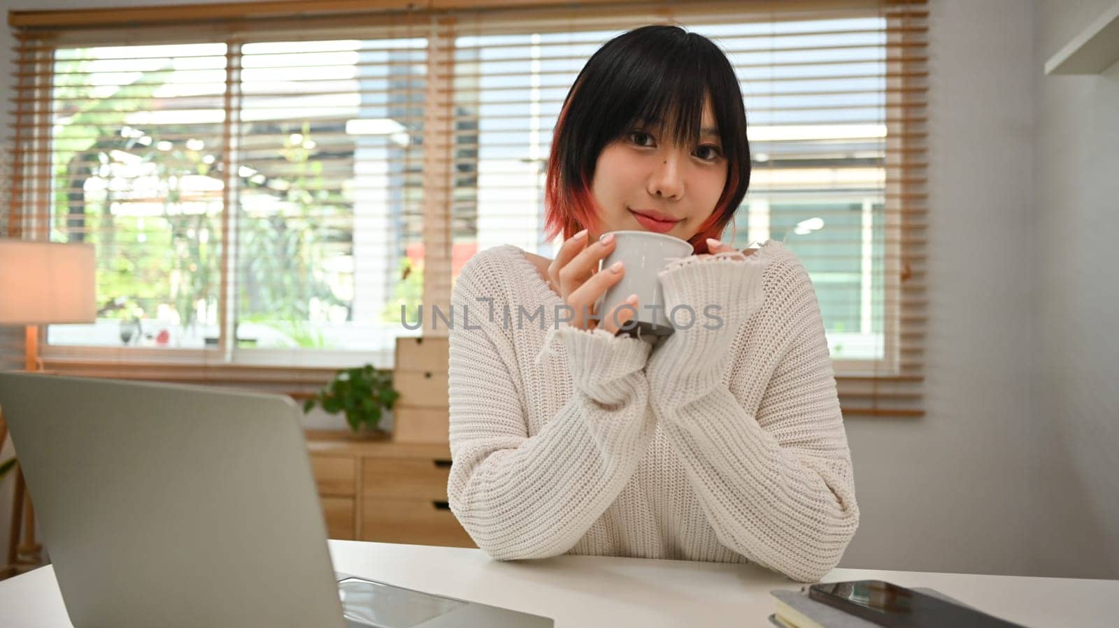 Pleasant young asian woman drinking coffee, sitting in front of laptop computer at home office desk.