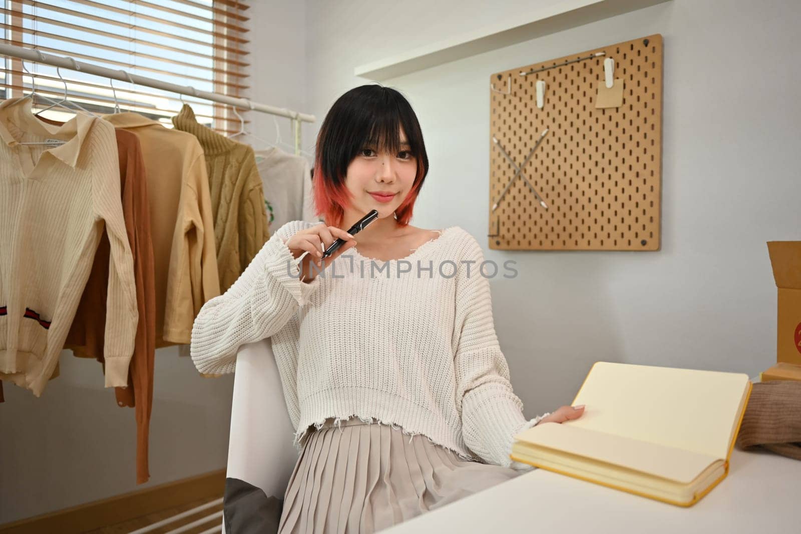 Young female fashion designer standing near clothing rack, working on new womenswear collection in modern studio.
