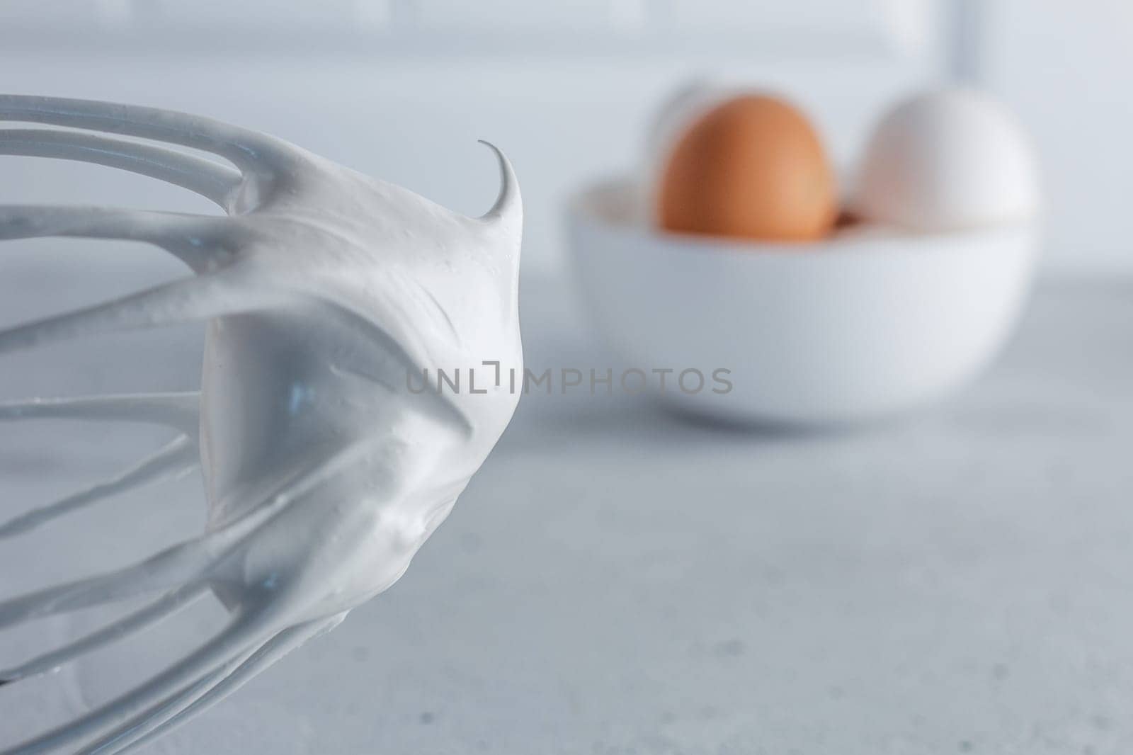 Whisked egg whites - whipped Italian meringue on a wire whisk and eggs on a gray background. copy space.