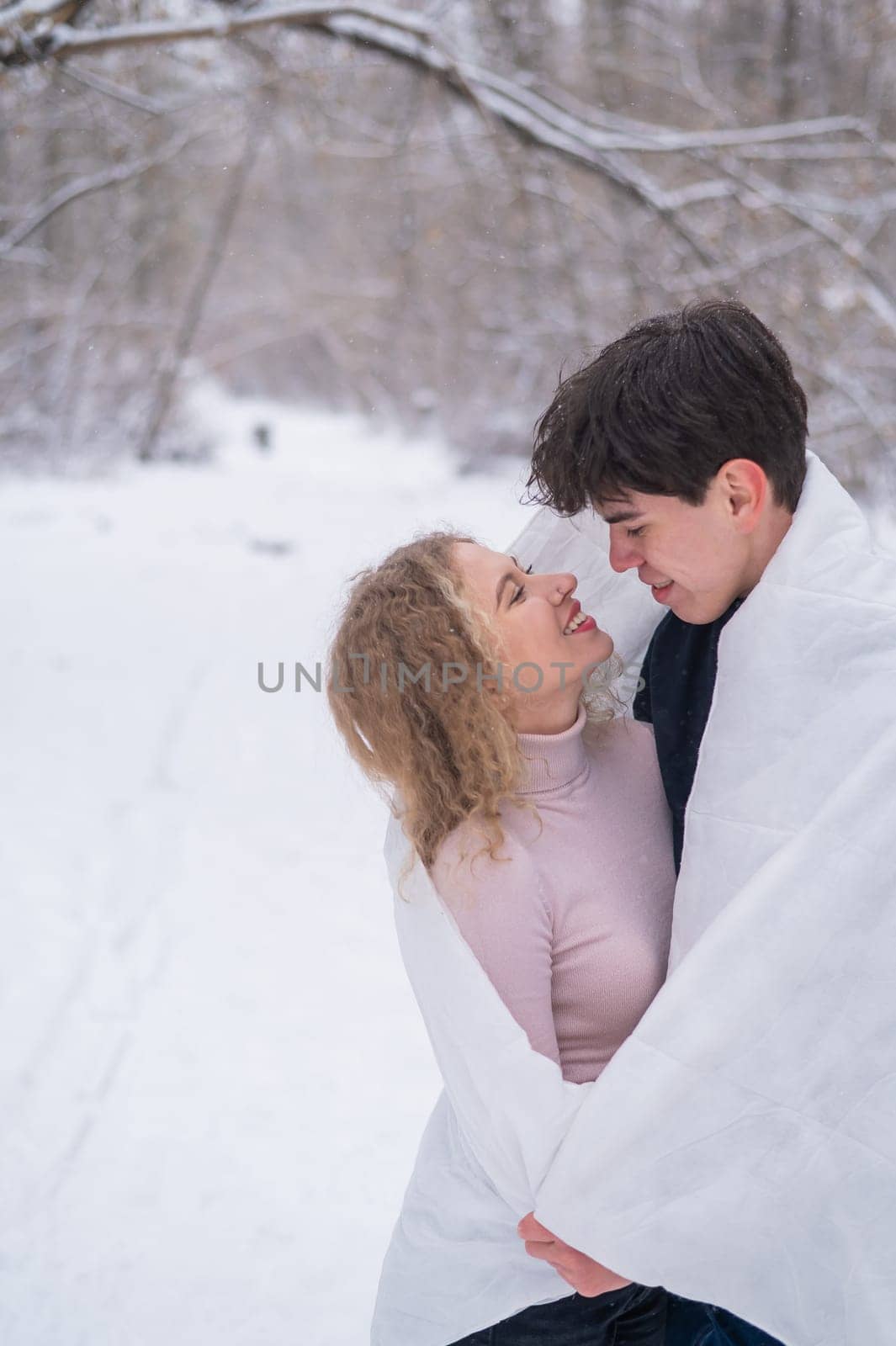 A young couple walks in the park in winter. The guy and the girl are kissing wrapped in a white blanket outdoors. by mrwed54