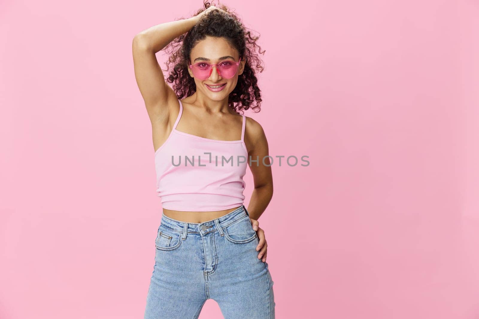 Happy woman hands up waxing her armpits, shaving, with curly hair in a pink tank top and jeans on a pink background wearing sunglasses with a nice tan, copy space. High quality photo