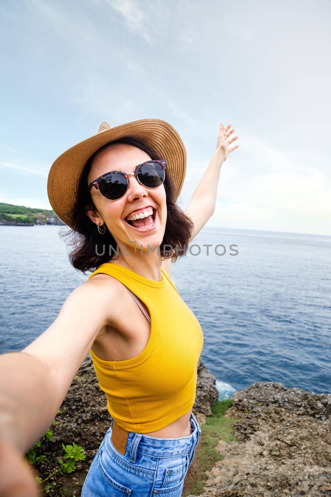 Joyful and smiling young woman traveler taking selfie with phone during summer vacation in seaside location. Vertical image. Travel, freedom and happiness concept.