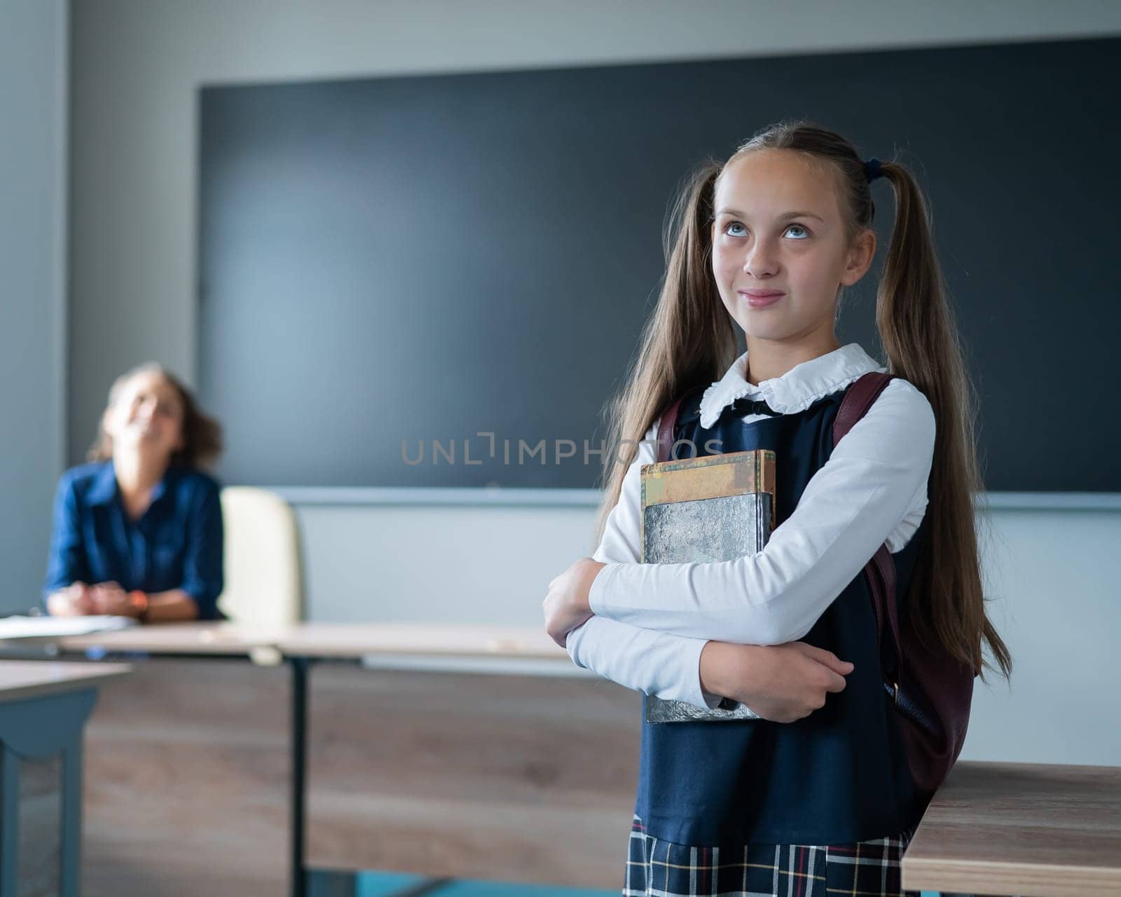 Caucasian girl and female teacher in the classroom. The schoolgirl is holding a textbook