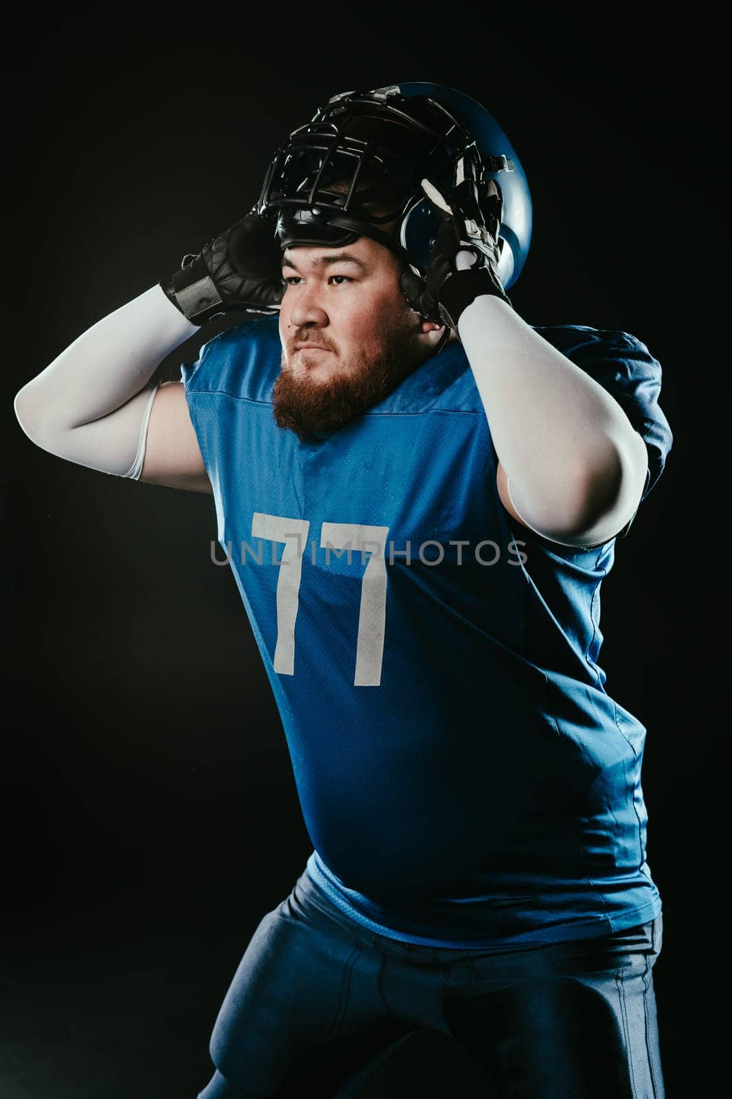 An Asian man with a red beard in a blue american football uniform puts on a helmet on a black background