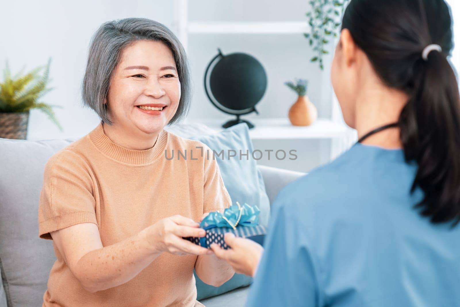 A young caregiver hand over to her senior patient a blue gift box with blue ribbons at a contented living room.