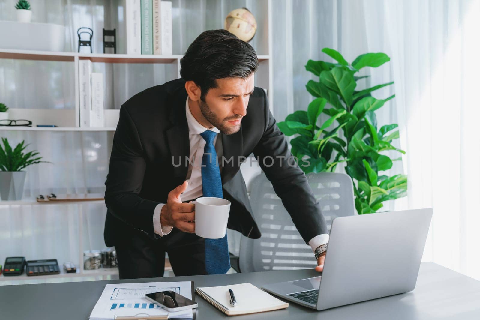 Modern professional businessman at modern office desk using laptop to work with coffee in his hand. Diligent office worker working on computer notebook in his office work space. fervent