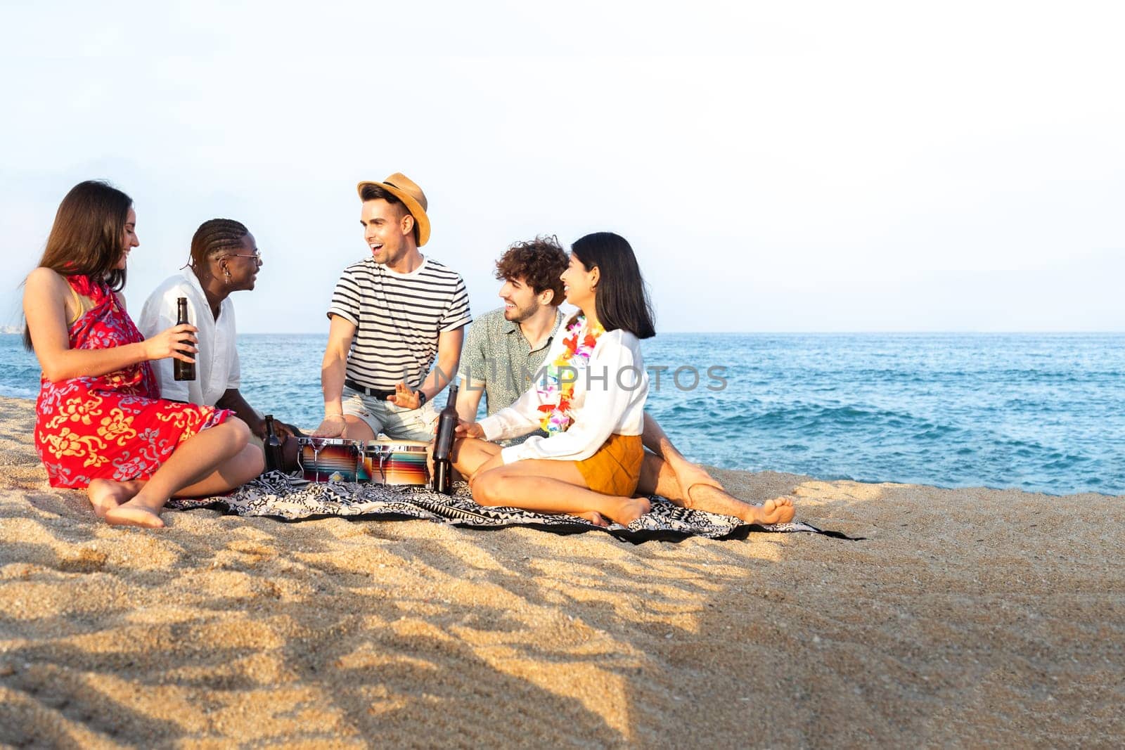 Group of multiracial having fun at the beach, talking, playing bongos and drinking beer together. Copy space. Vacation and friendship concept.