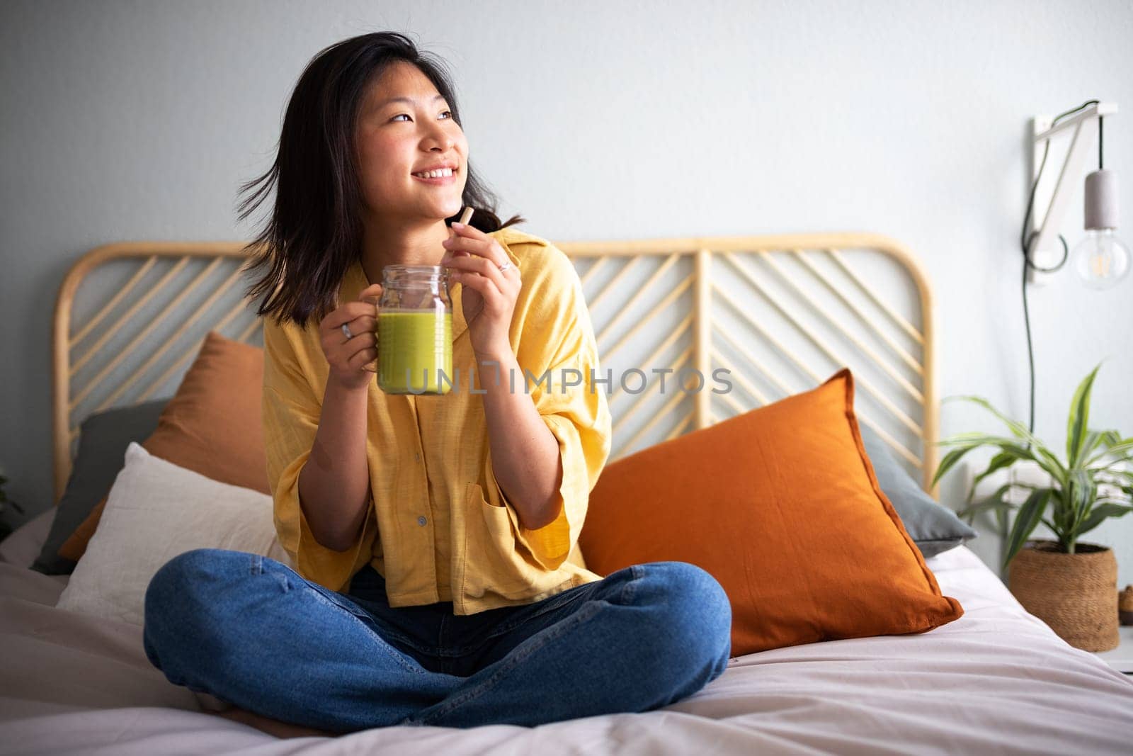 Pensive, happy young asian woman sitting on bed drinking green juice smoothie. Healthy lifestyle concept.