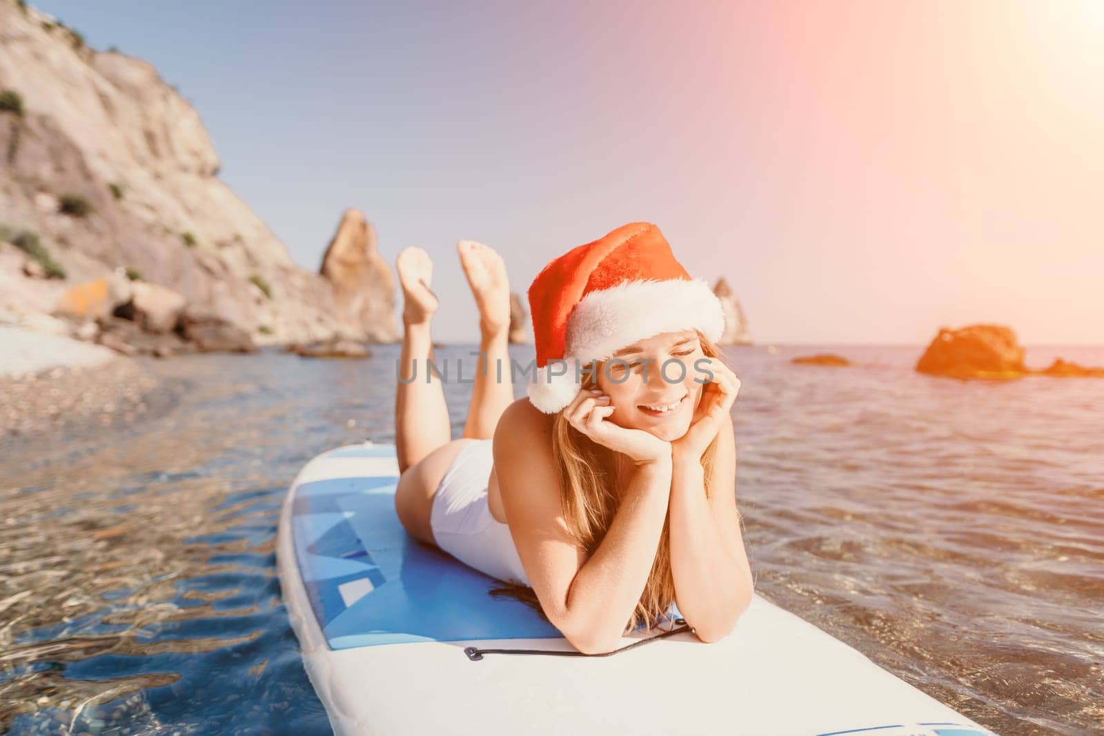 Woman sea sup. Close up portrait of happy young caucasian woman with long hair in Santa hat looking at camera and smiling. Cute woman portrait in a white bikini posing on sup board in the sea by panophotograph