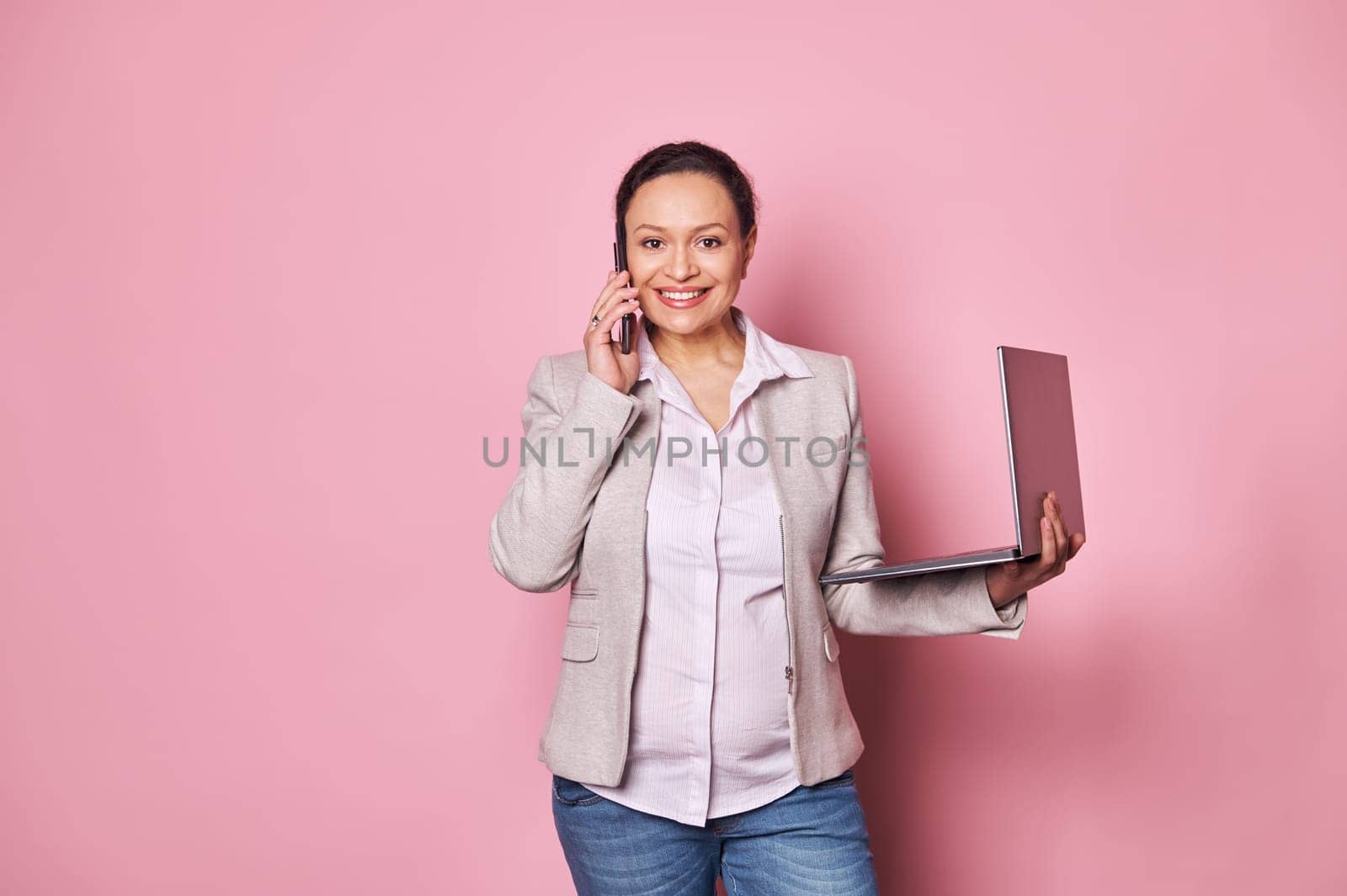 Stylish pregnant businesswoman talking on mobile phone, negotiating with business partners, posing with laptop on pink by artgf