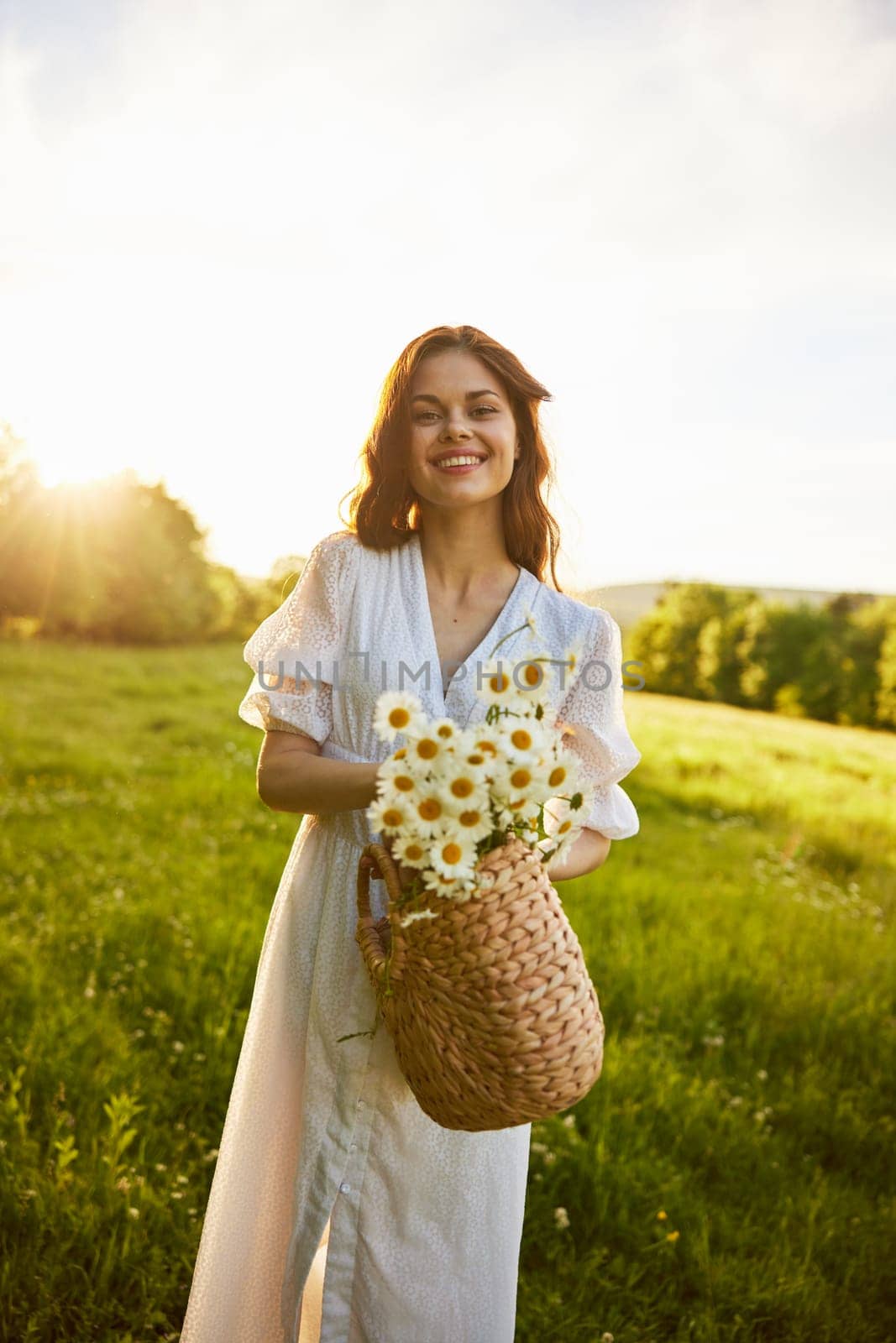 a woman in a light dress stands in a field during sunset and holds out a basket of daisies into the camera. High quality photo