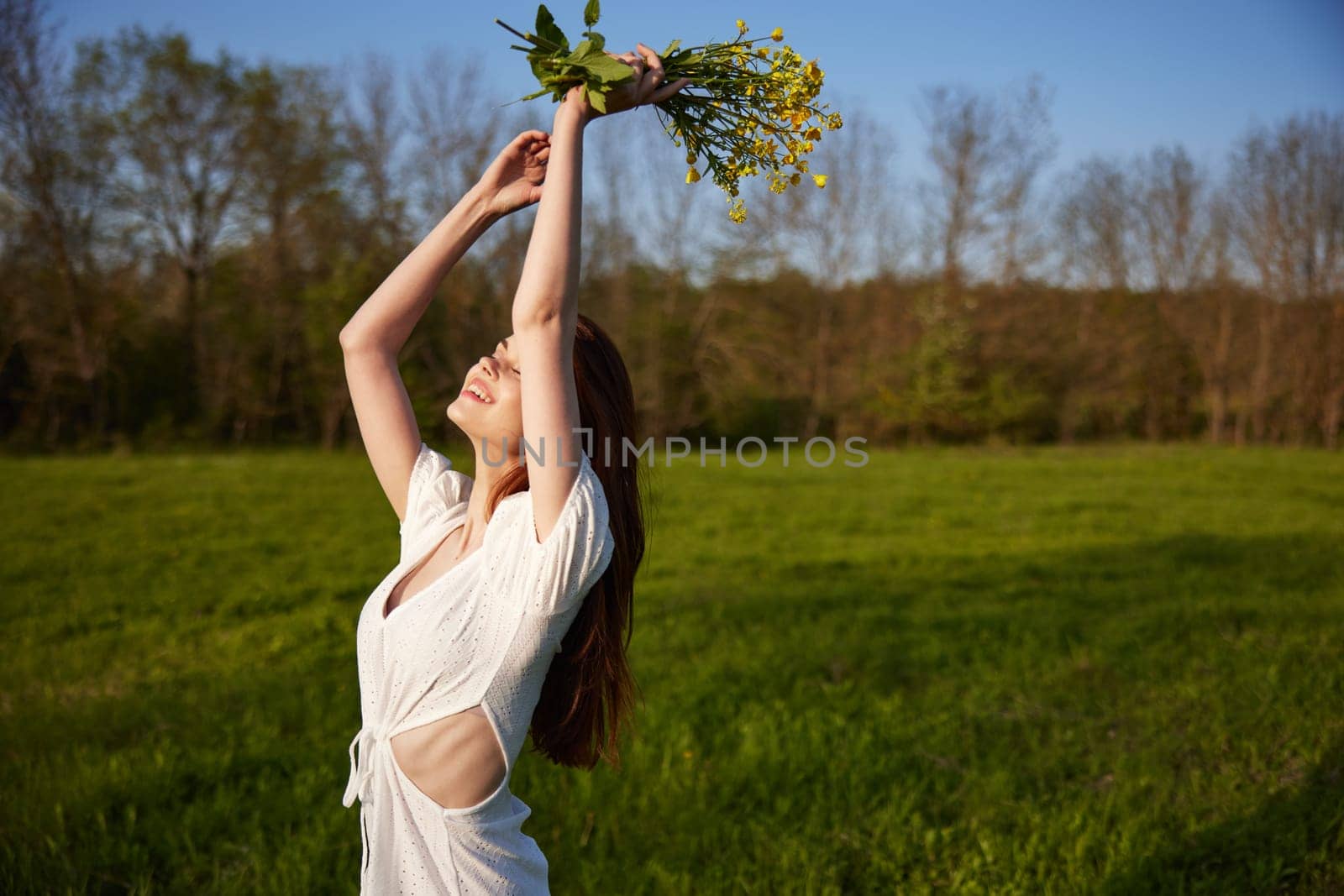 a beautiful, happy woman in a light dress stands in a field raising her hands high holding a bouquet of flowers. High quality photo
