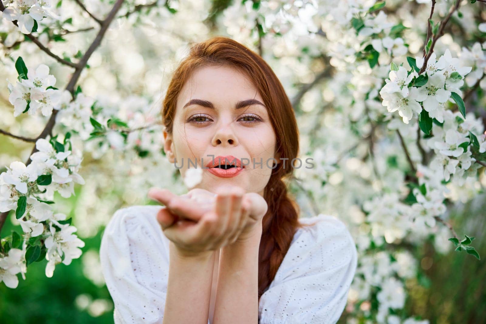portrait of a cute girl in a light dress against the backdrop of a blooming white tree blowing flowers from her palm into the camera. High quality photo