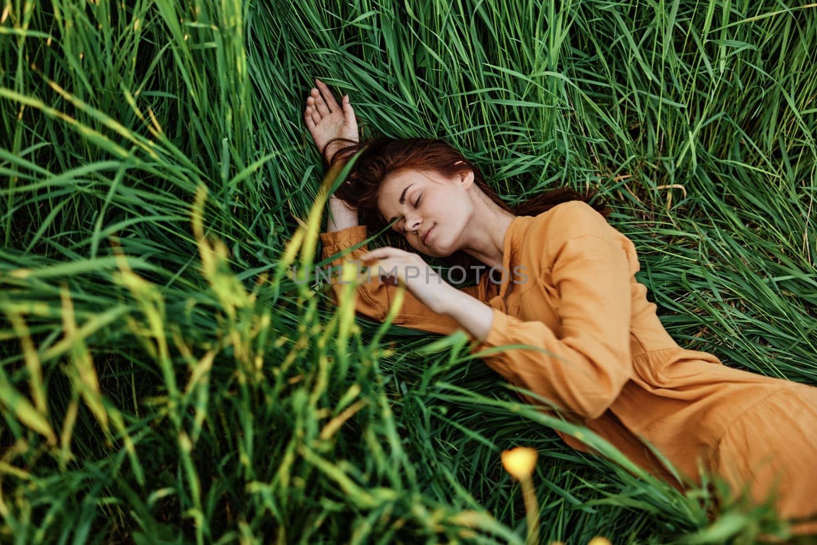 a close horizontal photo of a pleasant woman in a long orange dress resting lying in the tall grass with her eyes closed in sunny weather with her arms outstretched by Vichizh