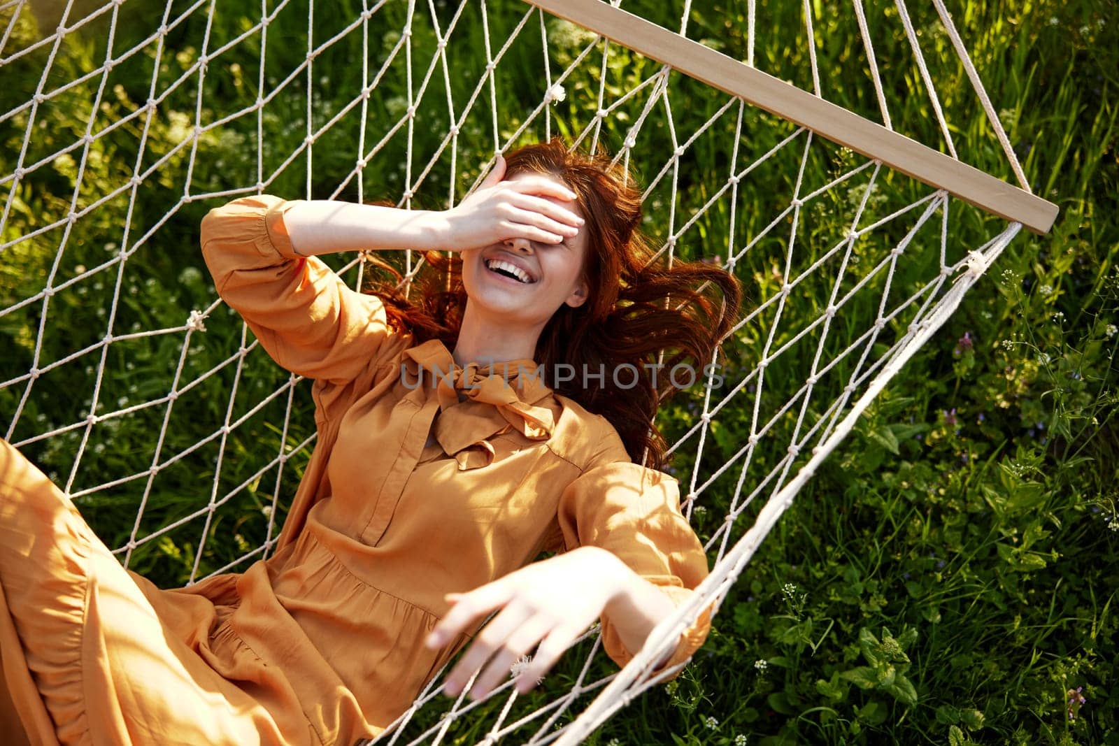 a beautiful, elegant woman lies in a long orange dress on a mesh hammock resting in nature, illuminated by the warm sunset light, covering her face with her hands. High quality photo