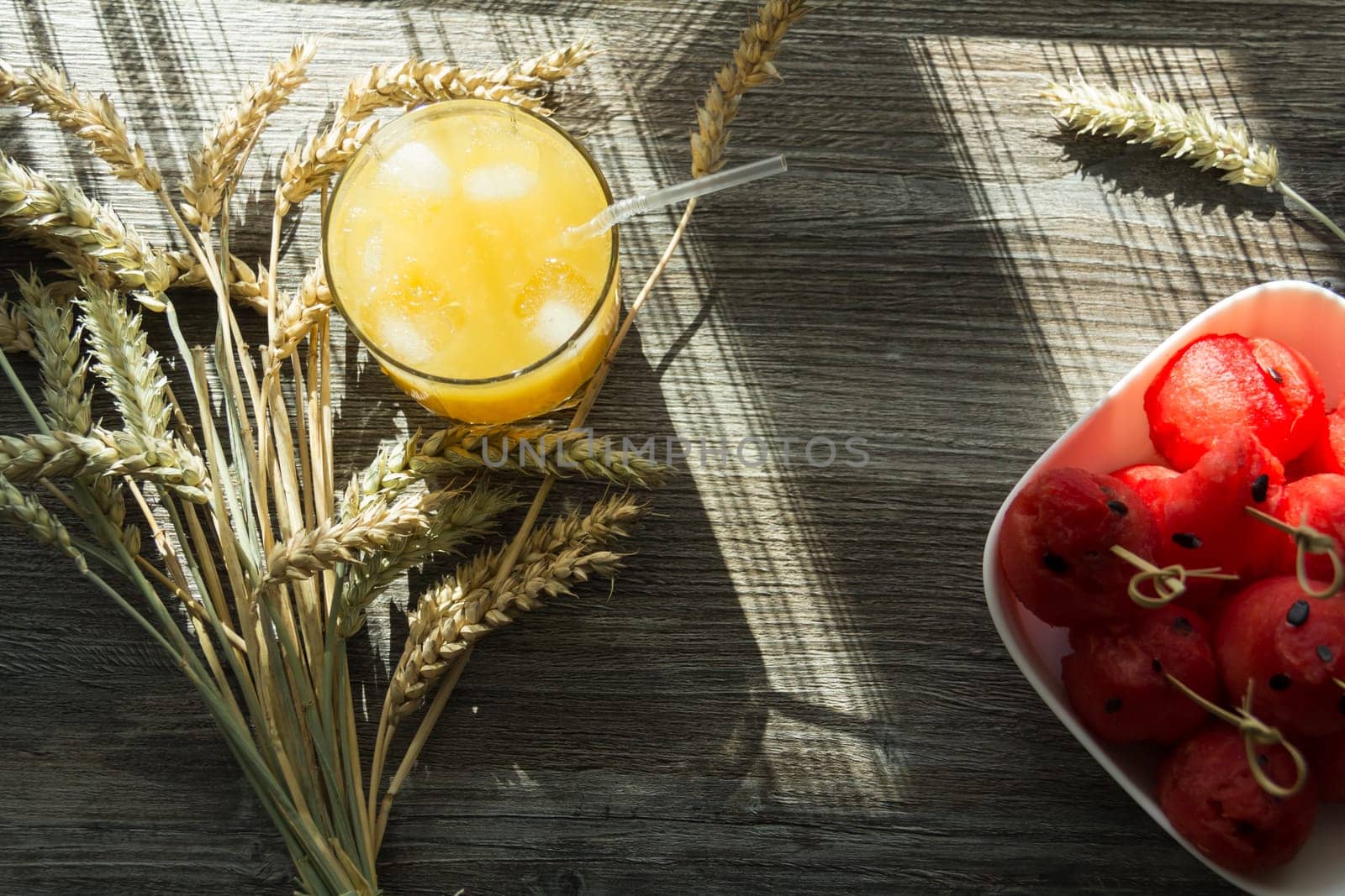 Freshly squeezed orange juice, sweet watermelon dessert and spikelets of ripe wheat on a wooden table in the sunlight