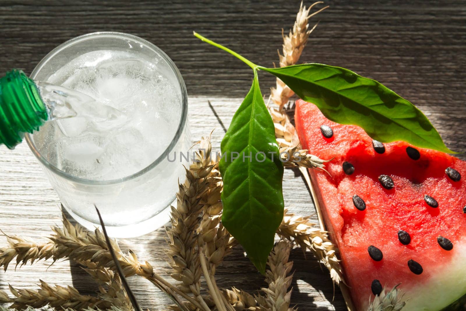 A triangular piece of sweet watermelon on an ice cream shelf, a cold drink with ice and ripe wheat ears on a wooden surface.