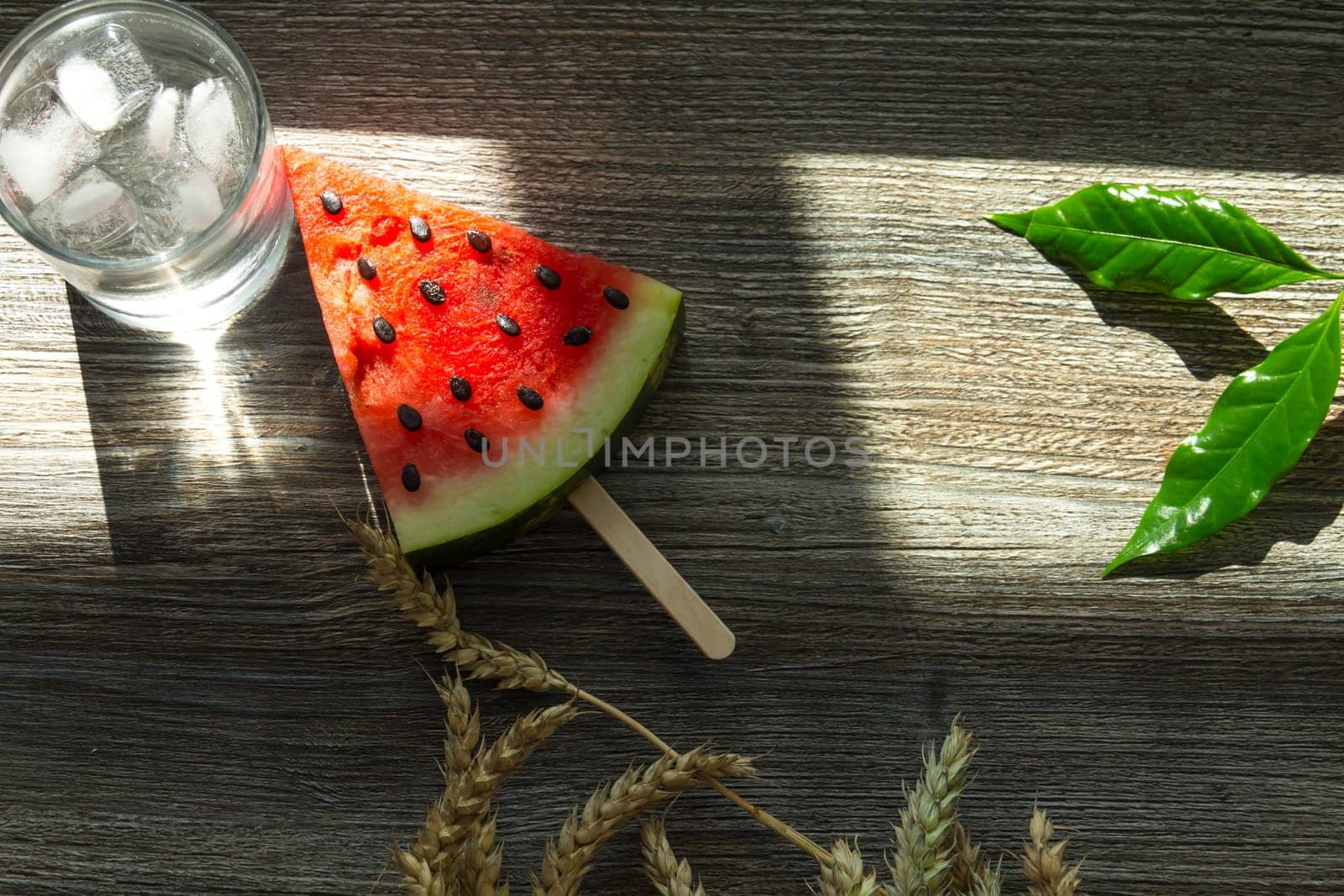 A triangular piece of sweet watermelon on an ice cream shelf, a cold drink with ice and ripe wheat ears on a wooden surface.,,