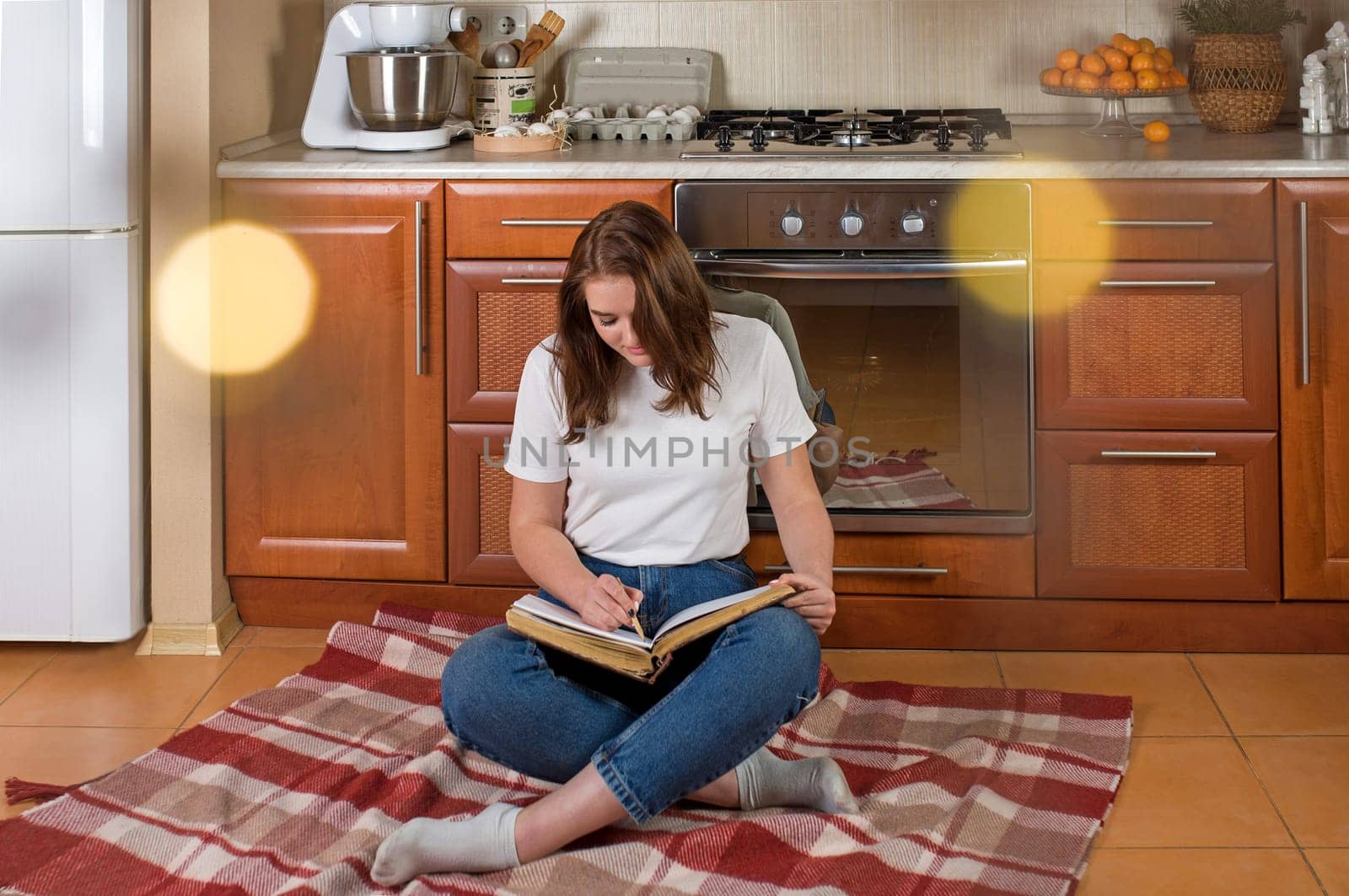 Horizontal shot of a happy woman sitting on the floor in the kitchen with an open notepad waiting for a delicious pie baking in the oven by aprilphoto