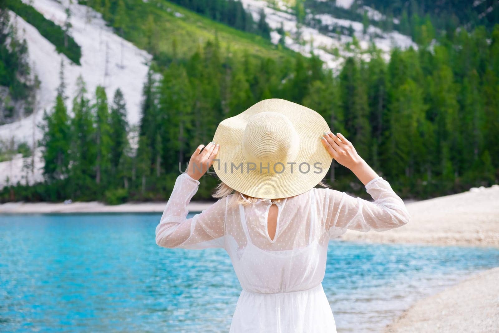 Young girl enjoying beauty of nature looking at mountain and lake Braies. Travel to Italy. A woman stands with raised arms and holds a summer hat on her head.