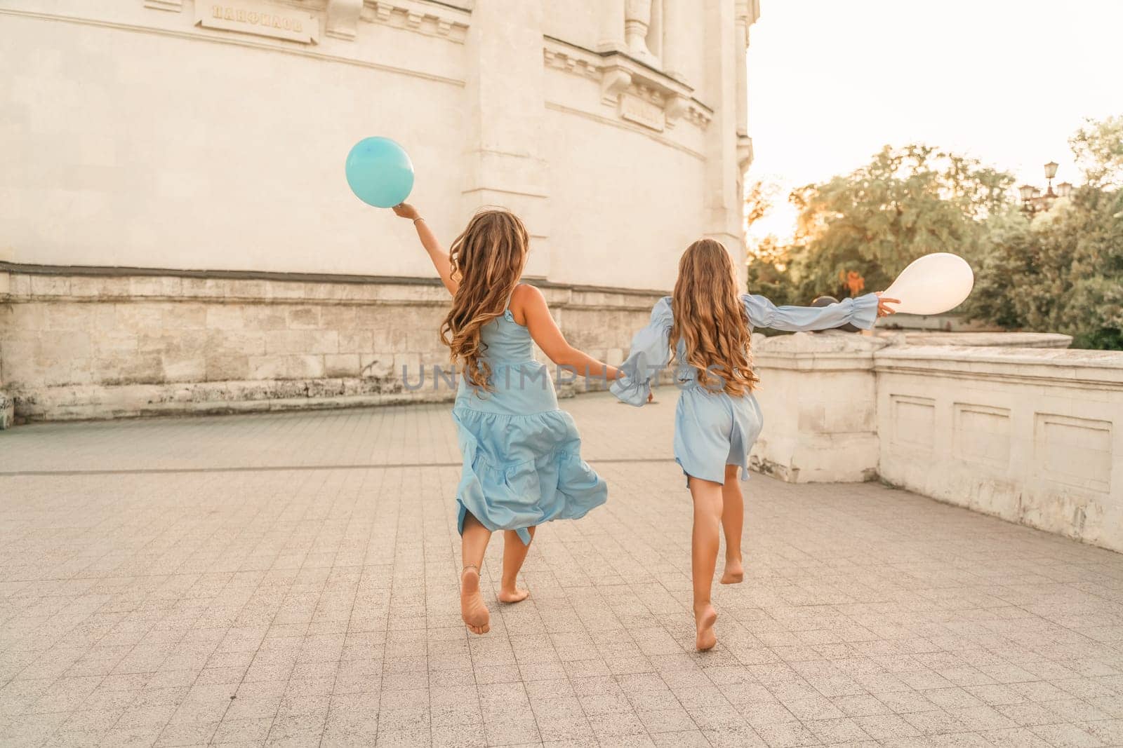 Daughter mother run holding hands. In blue dresses with flowing long hair, they hold balloons in their hands against the backdrop of a sunset and a white building. by Matiunina