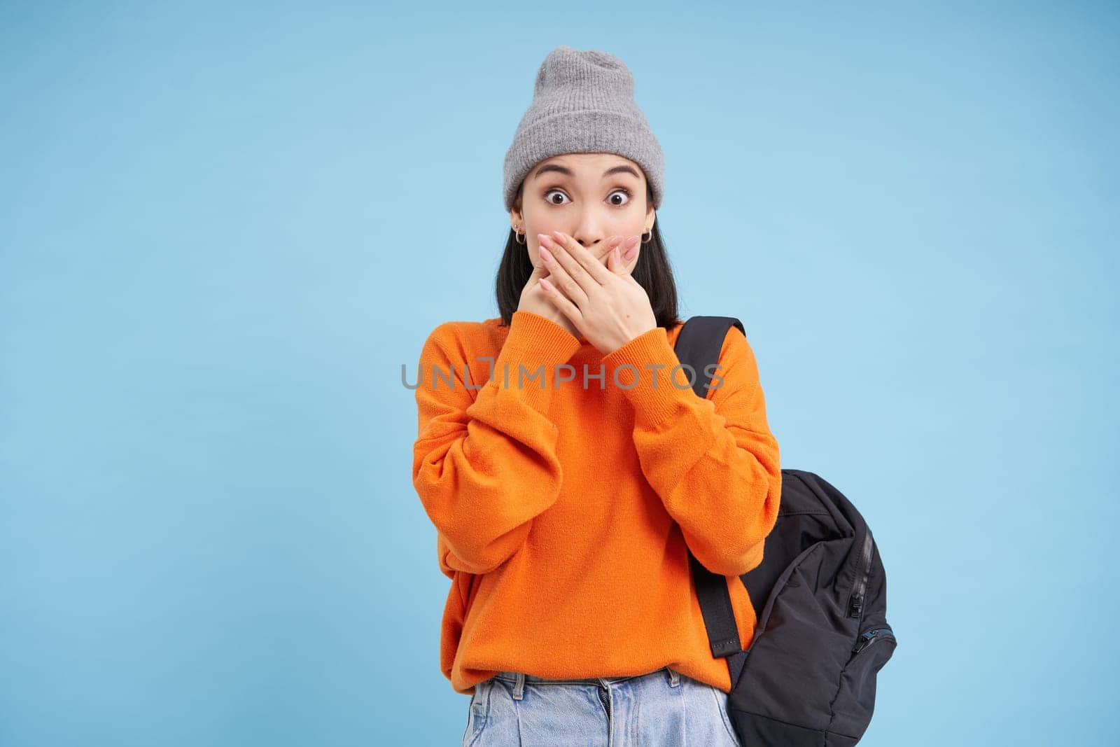 Portrait of korean girl in hat, covers her mouth with hands, looks shocked, amazed by gossips, stands over blue background.