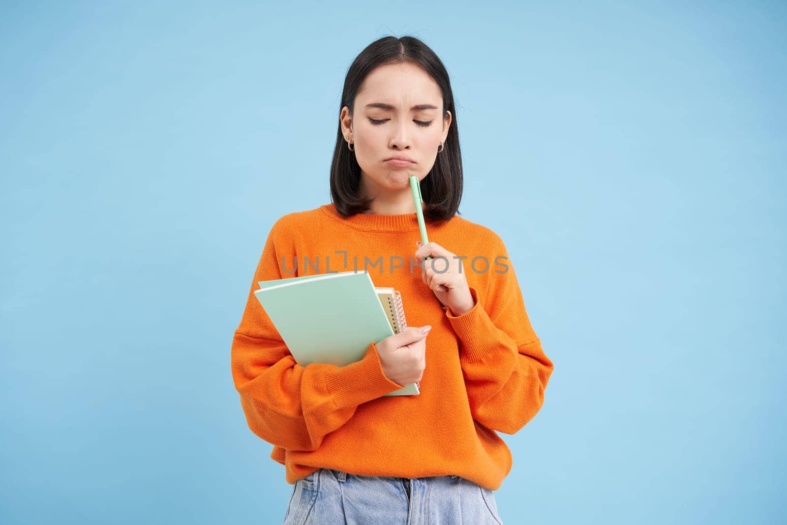 Education and students. Happy asian woman, holding notebooks and laughing, smiling at camera, enjoys going to University or College, blue background.