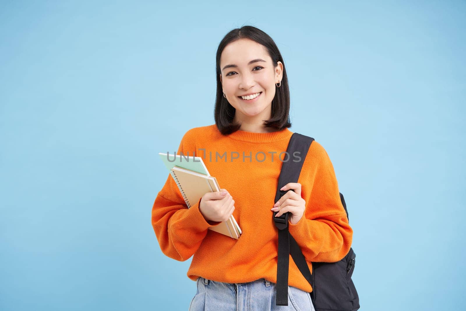 Education and students. Smiling young asian woman with backpack and notebooks, posing against blue background by Benzoix