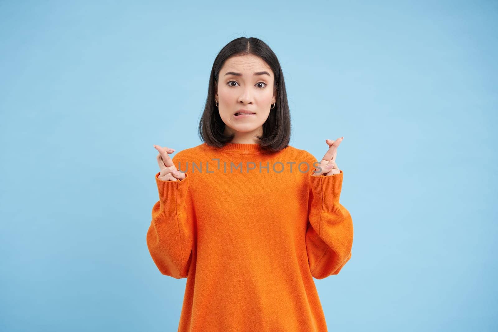 Portrait of asian woman cross fingers, hopes and prays for something, makes a wish, stands over blue background.