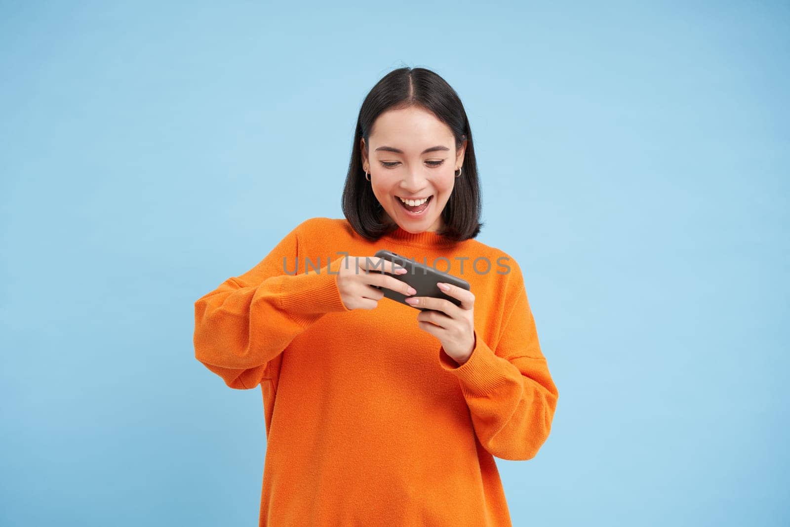 Happy smiling girl, 25 years, plays mobile video games, holds smartphone in both hands, stands against blue background.