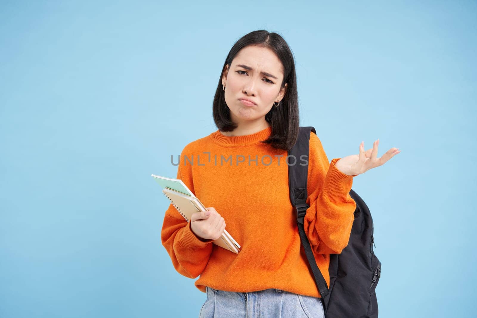 Annoyed young asian woman, student complans, shakes hand and looks disappointed, stands with backpack and notebooks, blue background.