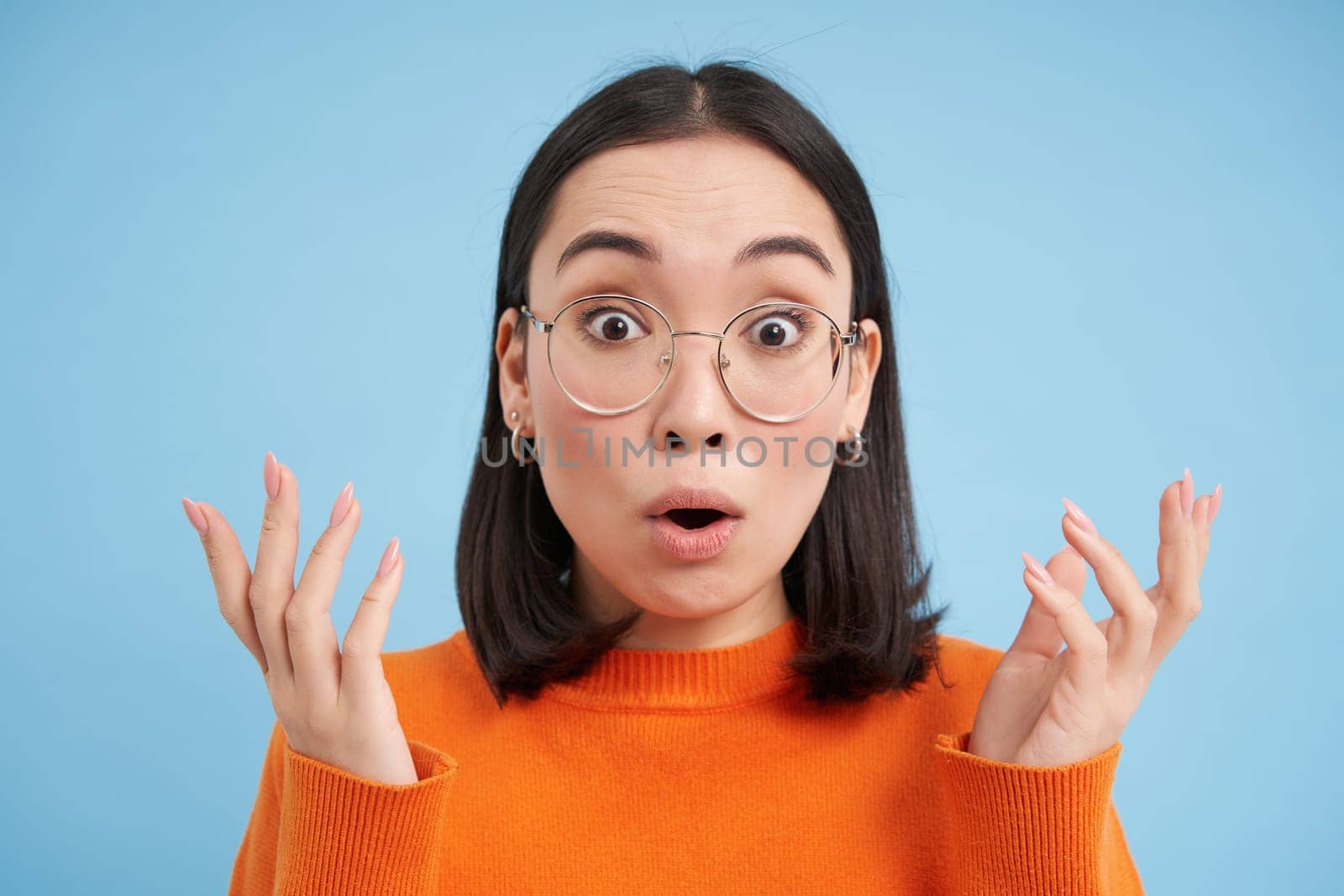 Glasses and eyewear, opticians concept. Portrait of beautiful asian woman in new spectacles, looks surprised, stands over blue background.