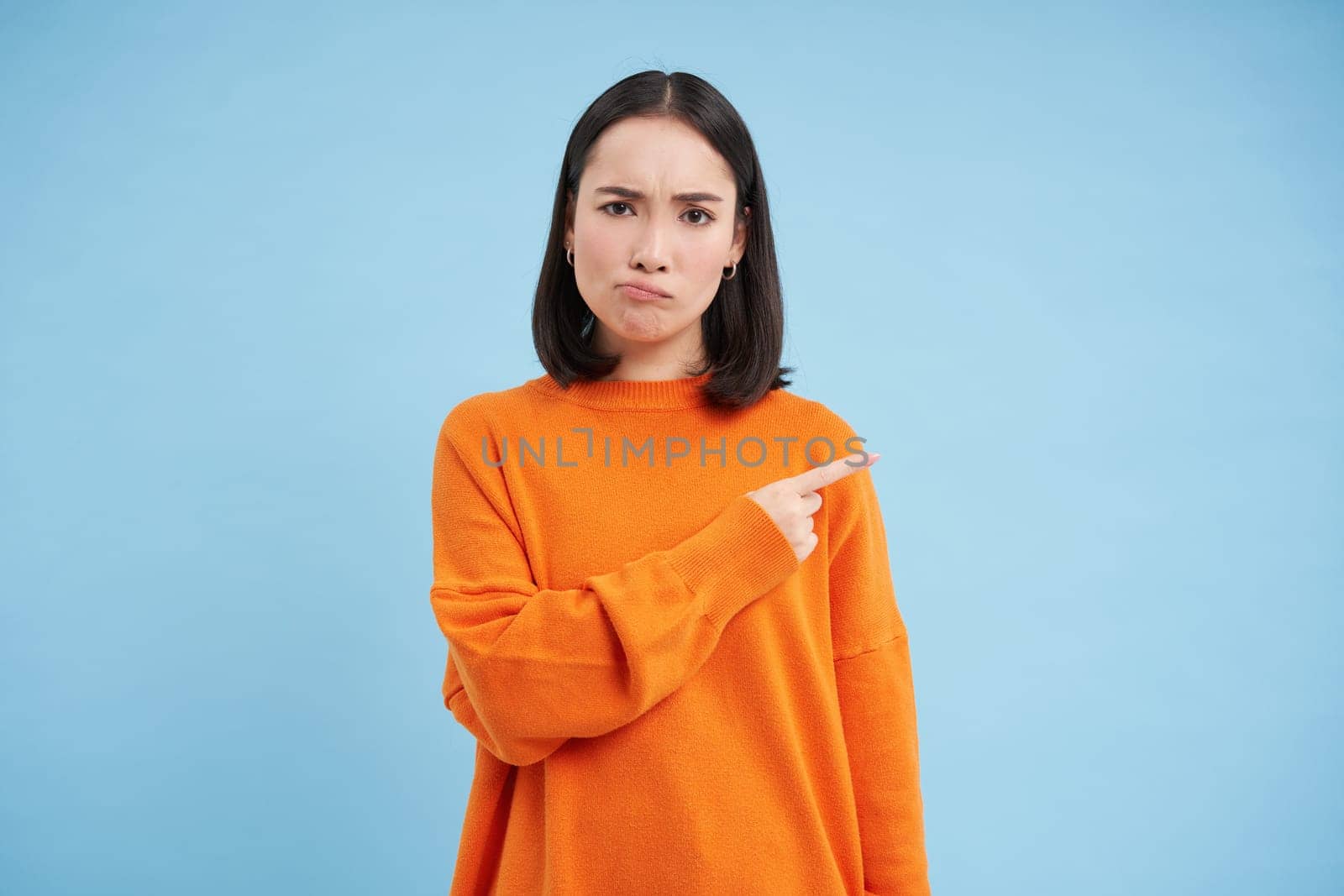 Upset brunette girl 25 years old, points right at banner, copy space on blue background, frownd with disappointed face expression by Benzoix