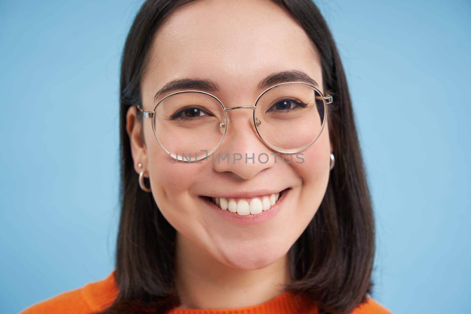 Close up portrait of beautiful young asian woman in glasses, smiling and looking happy, trying new eyewear at opticians store, stands over blue background by Benzoix