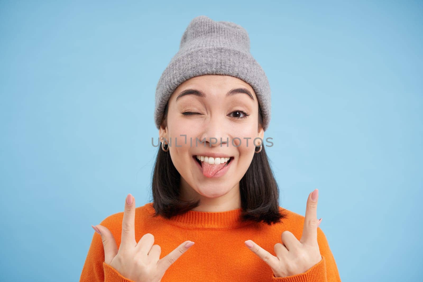 Close up portrait of funny and carefree asian girl in beanie, shows rock n roll, heavy metal gesture and tongue, stands over blue background.