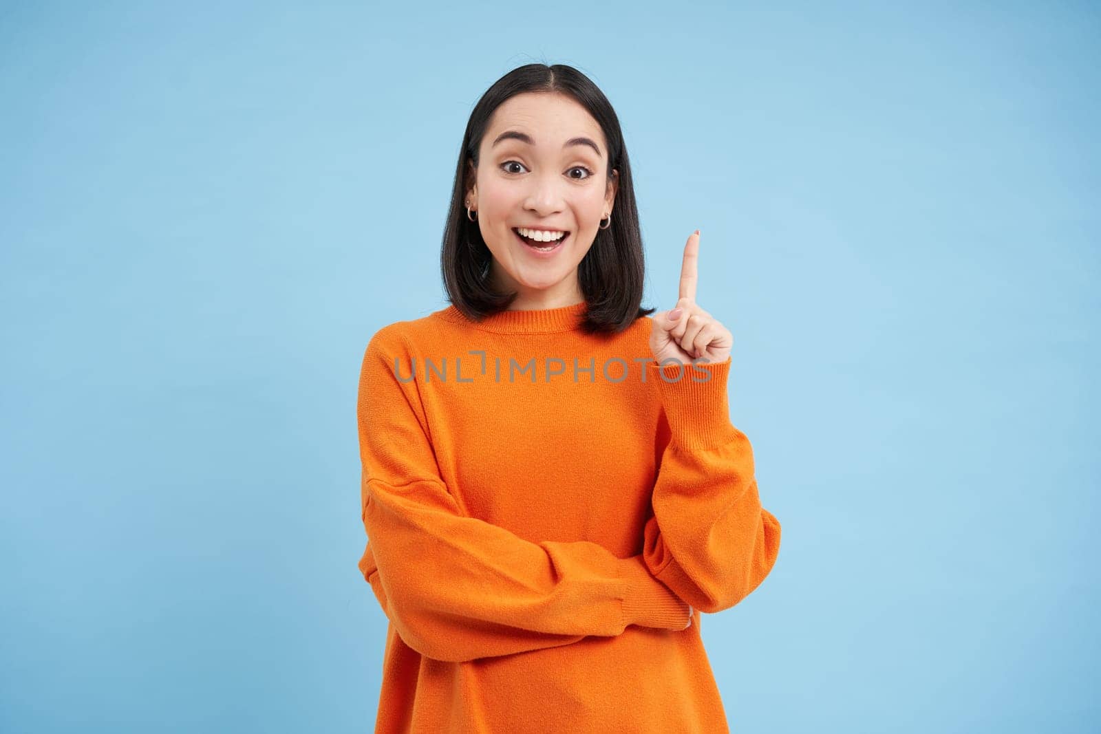 She has perfect plan. Amazed korean woman shows one finger, eureka gesture, has an idea, suggest something, say her thoughts, stands over blue background.