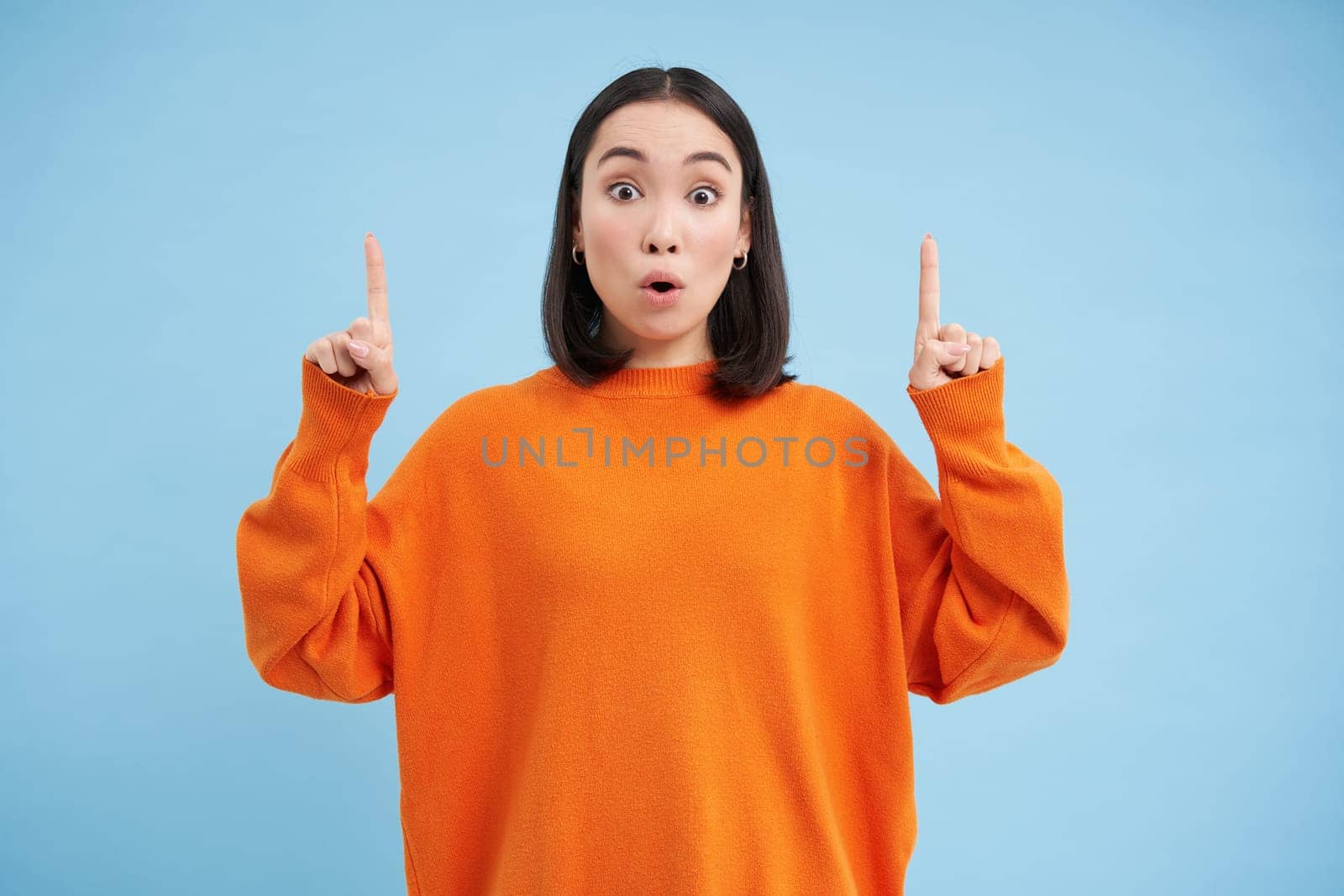 Amazed asian female model, says wow, looks impressed, points fingers up, stands over blue background by Benzoix