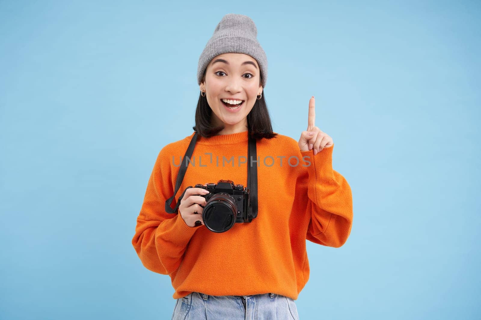 Surprised asian girl in hat, holds digital camera, looks amazed and impressed, poses over blue studio background with space for banner.