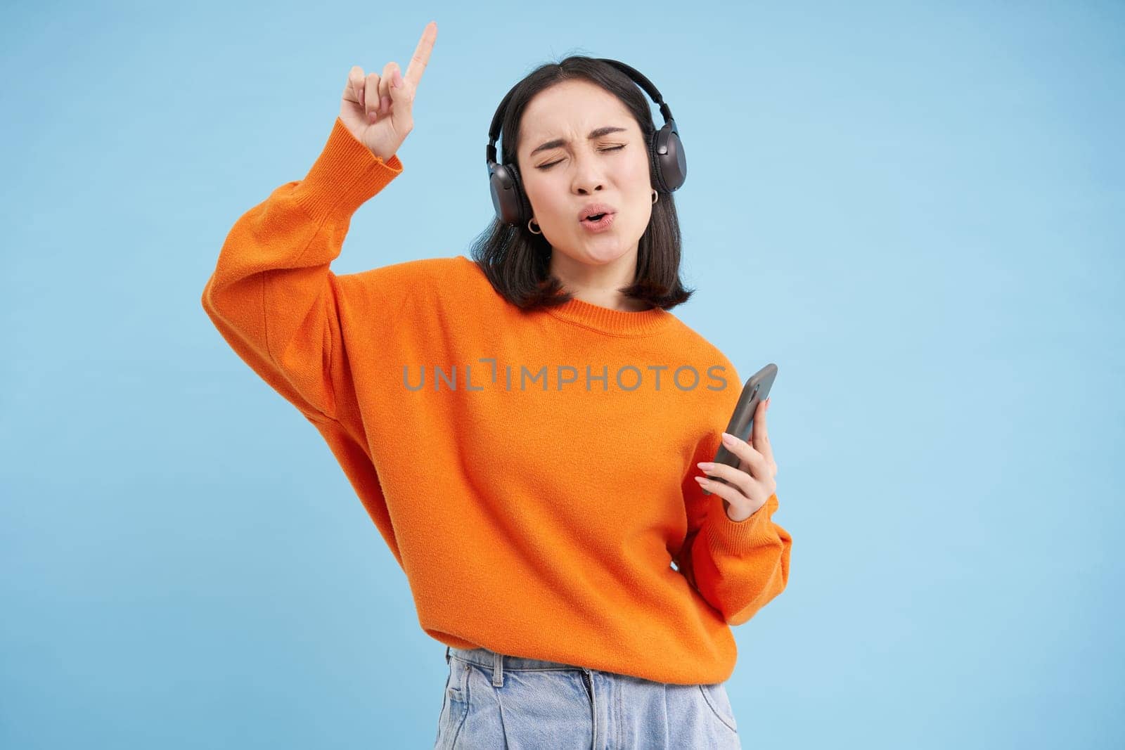 Beautiful korean woman in headphones, dancing, listening music on mobile phone app, smiling, posing over blue background.
