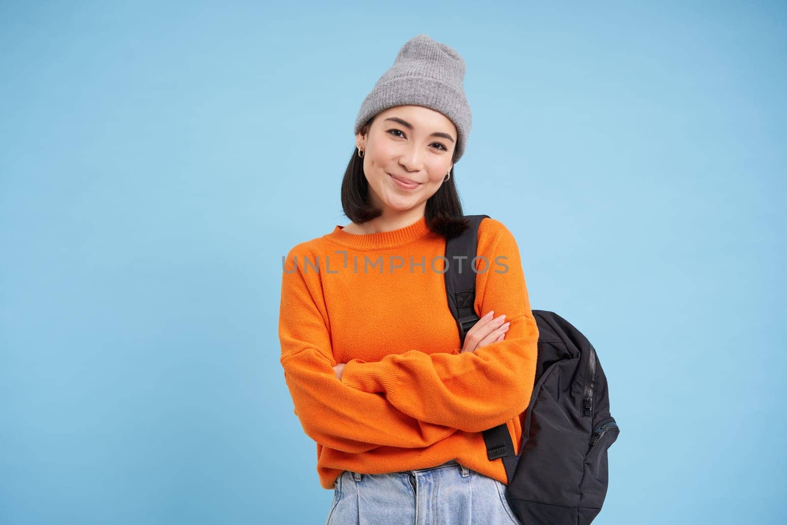 Cheerful chinese woman in warm hat and orange sweater, goes for walk with backpack, cross arms on chest and smiles with confidence, blue background.