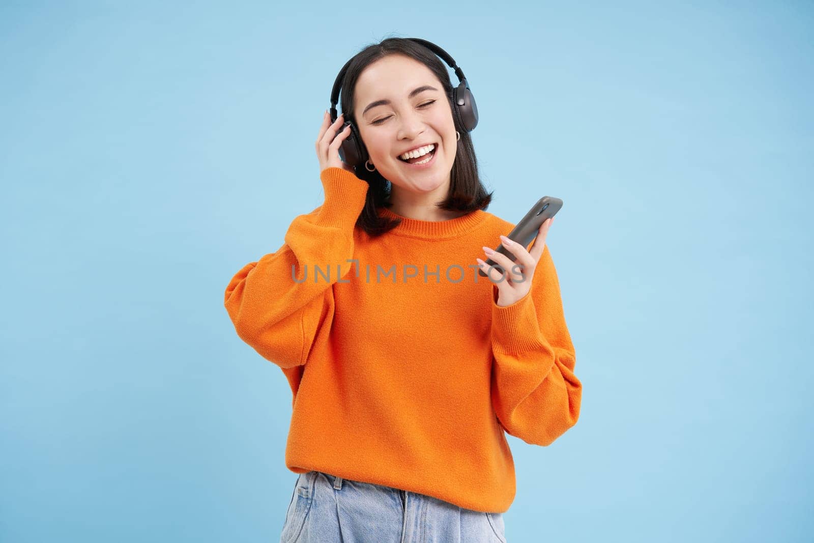 Young woman in headphones and with smartphone listens music, enjoys singing favourite songs in her playlist, blue background. Copy space
