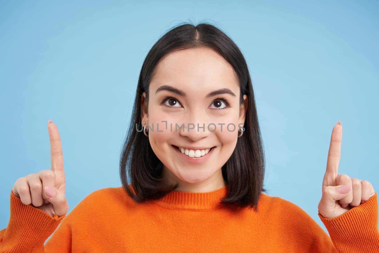 Beautiful asian girl points fingers up, shows banner on top, advertisement, stands over blue background. Copy space