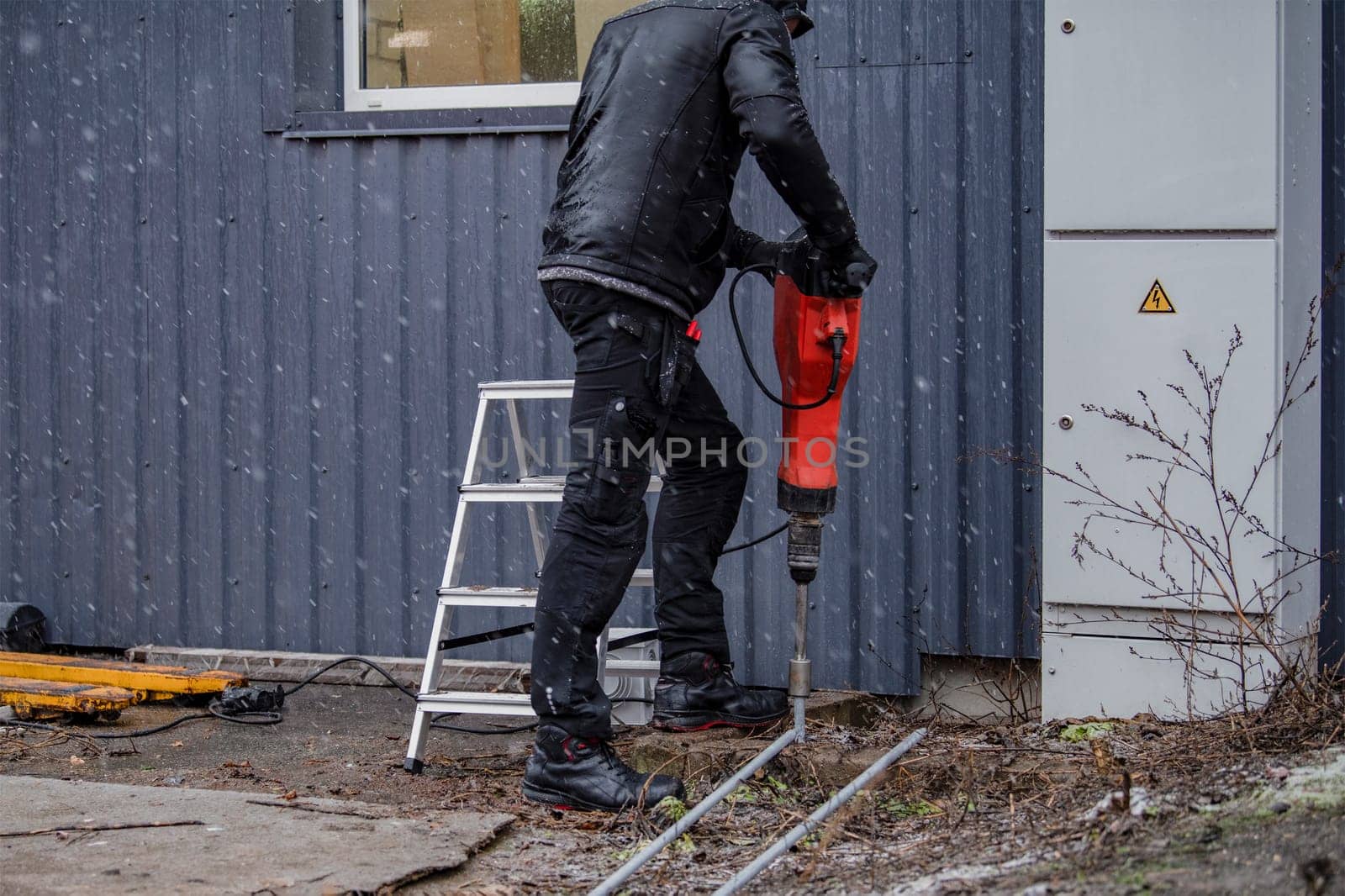 A worker installs a ground rod to ground a building. A worker in work clothes drives earth rods into the ground with a jackhammer to prevent short circuits