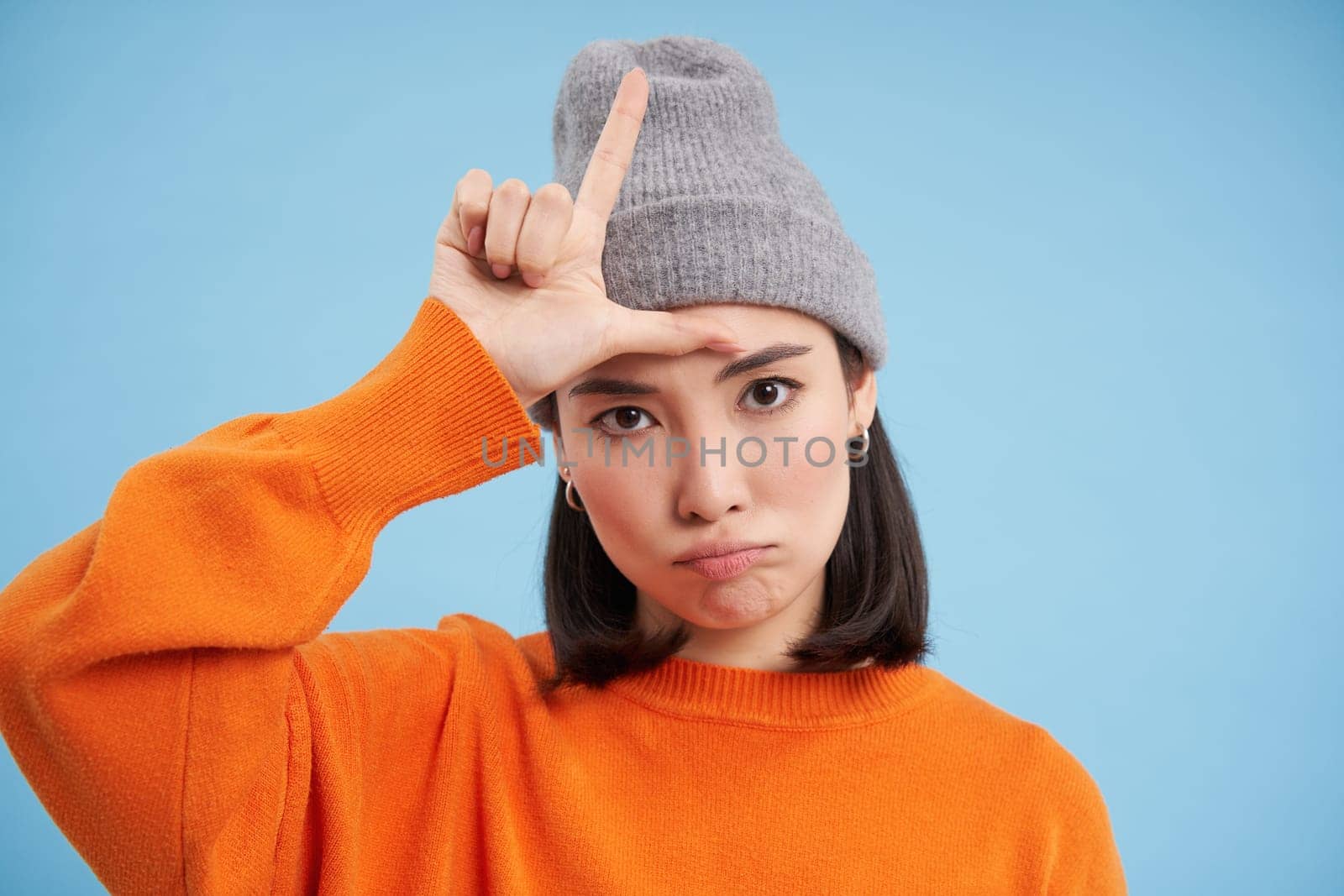 Close up portrait of asian girl student in beanie, shows L letter, loser sign on forehead, stands over blue background.